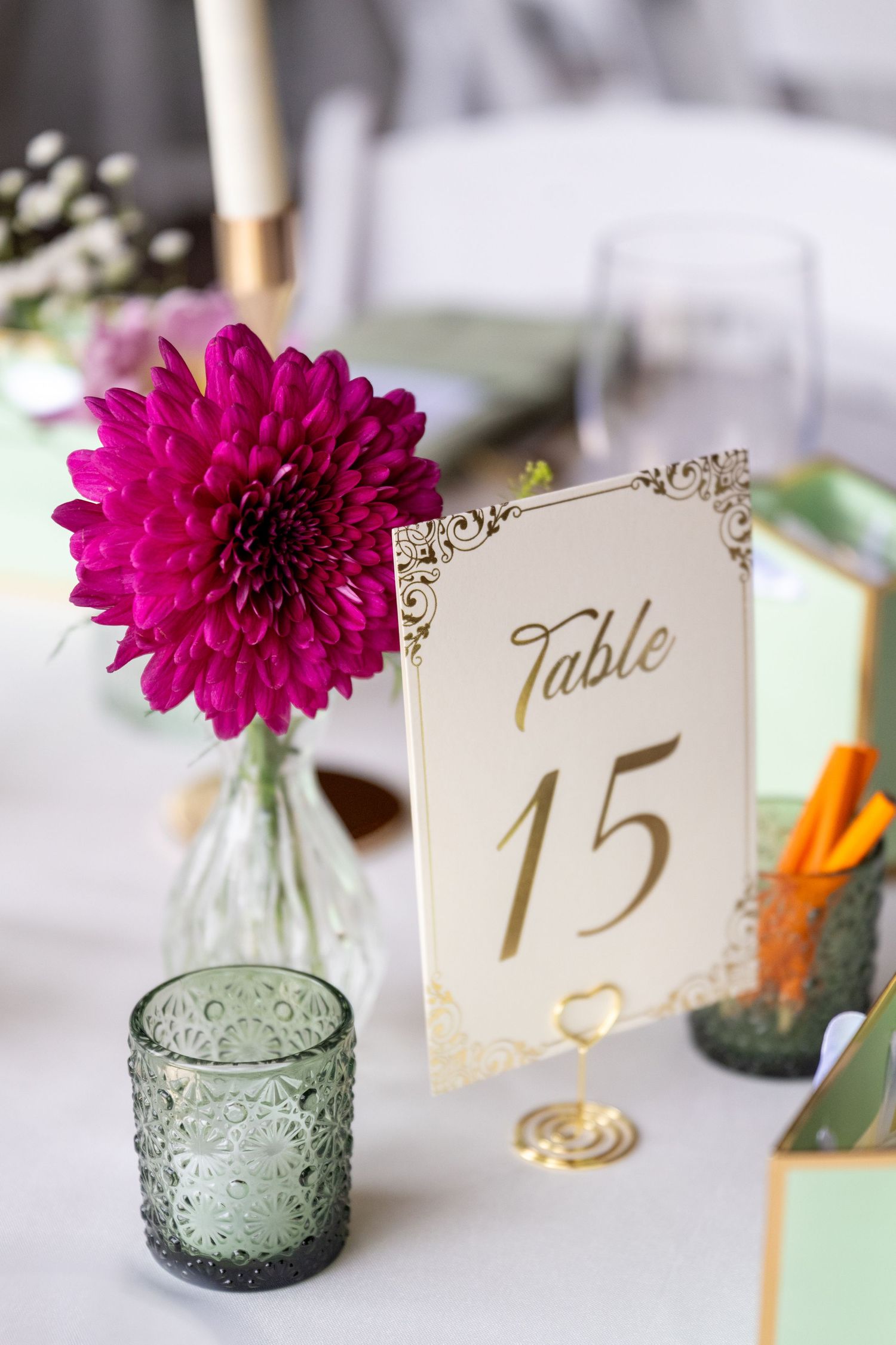 Elegant wedding table 15 sign in gold holder with vibrant pink dahlia in glass vase and green textured candle holder.