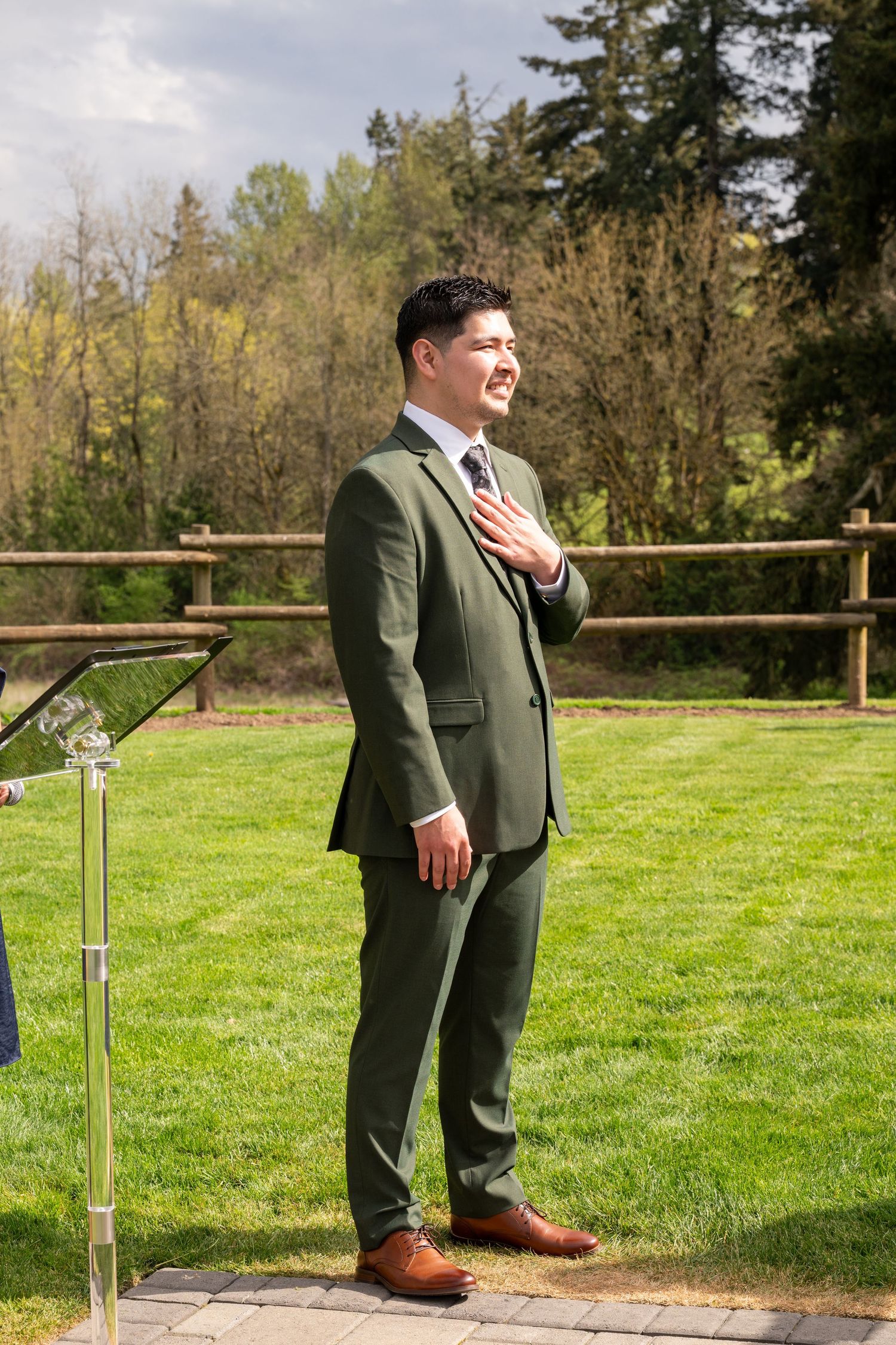 Groom in olive green suit with hand on chest, standing outdoors at wedding ceremony on sunny day.