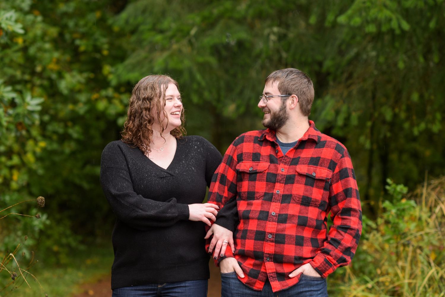 A couple shares a joyful moment together on a wooded path wearing coordinated red plaid and black outfits.