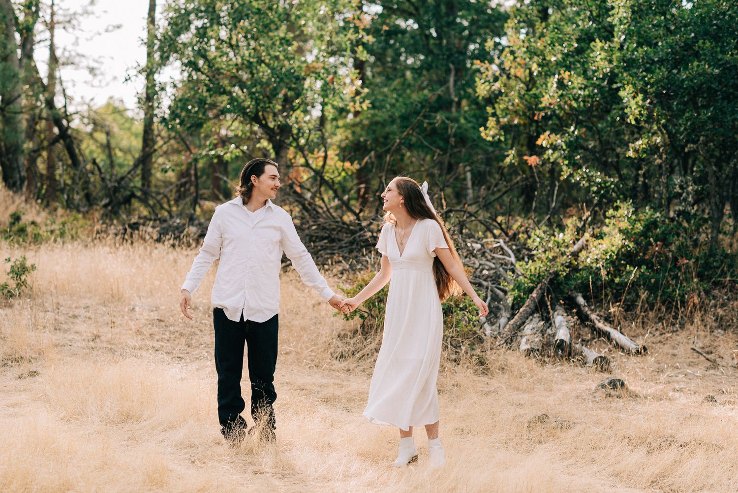 A couple in white clothing walking hand in hand through a sun-drenched field with trees in the background.