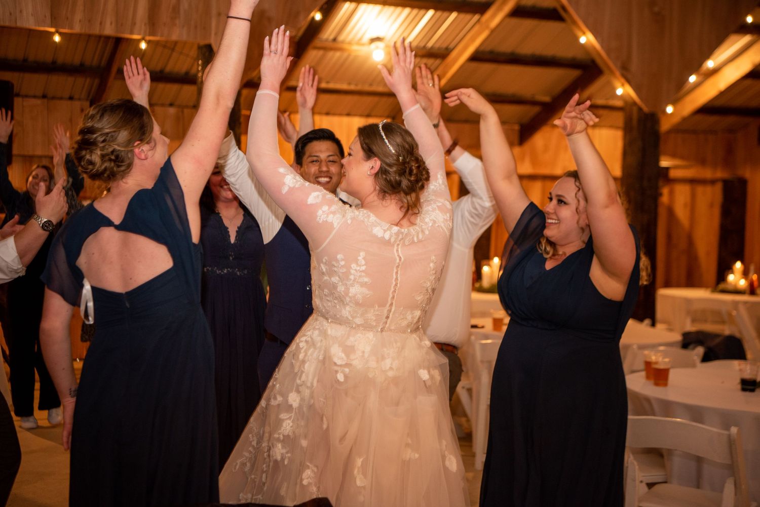 Wedding reception guests dance and celebrate on a wooden dance floor under warm lighting.