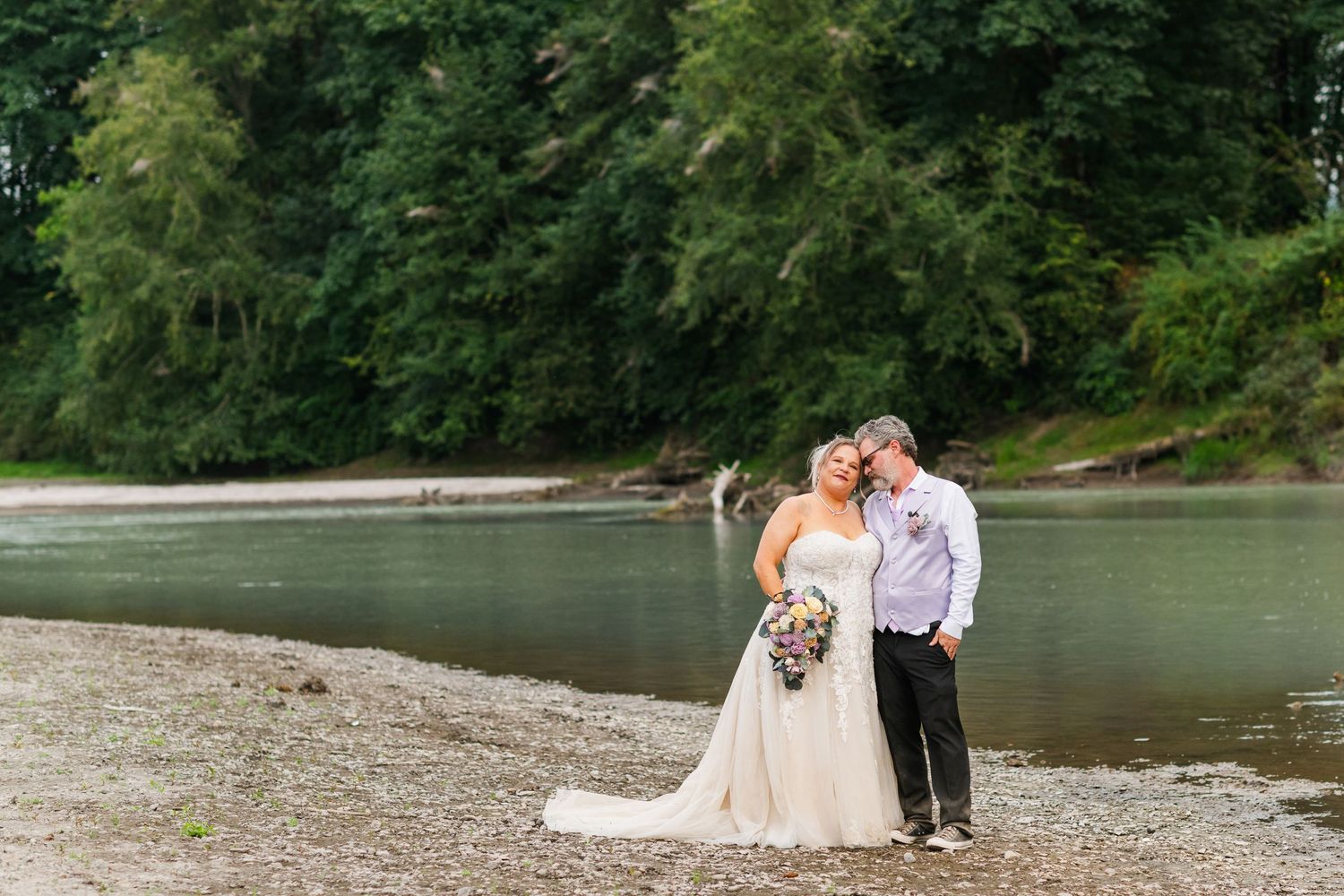 Wedding couple poses on a rocky riverbank surrounded by lush green trees.