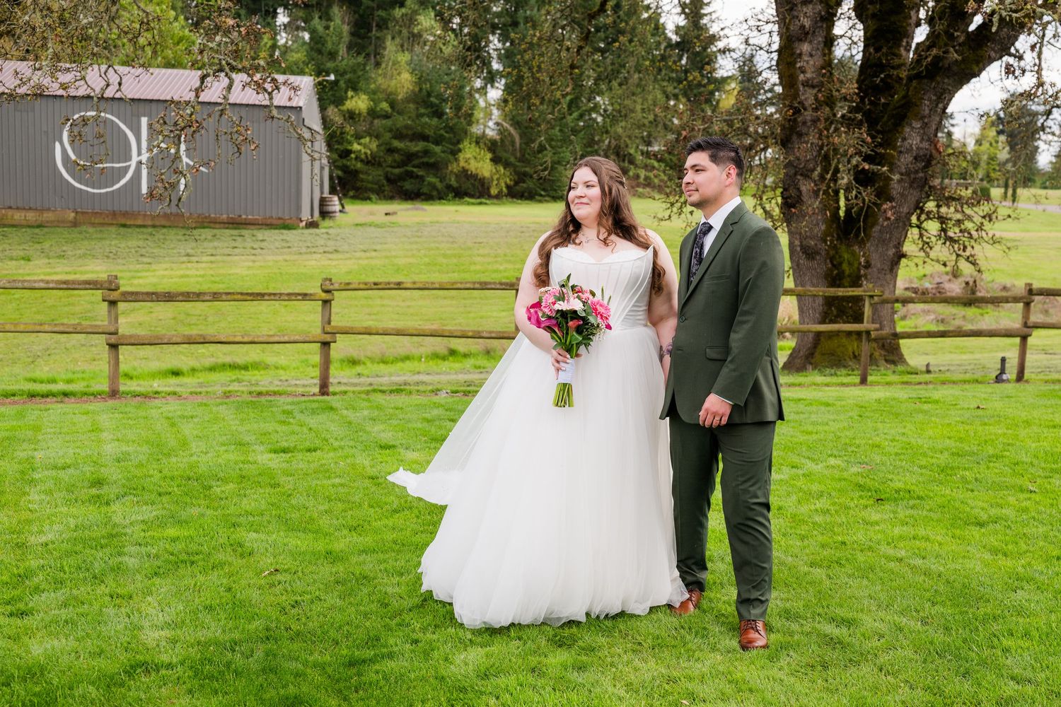 Bride in flowing white gown and groom in olive suit walk hand-in-hand across a lush green lawn at their wedding.