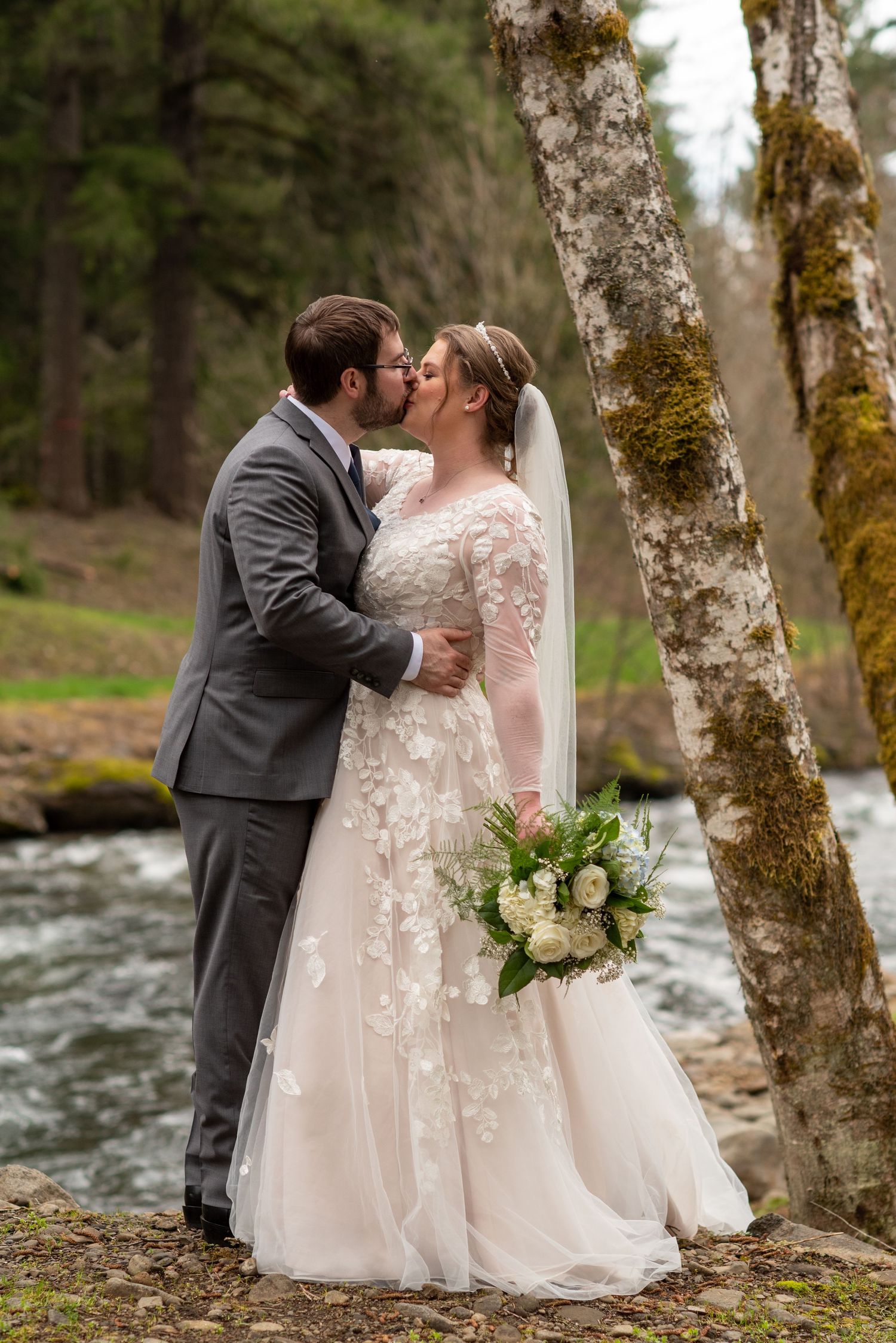 Bride and groom kissing by a forest river, bride holding white floral bouquet.