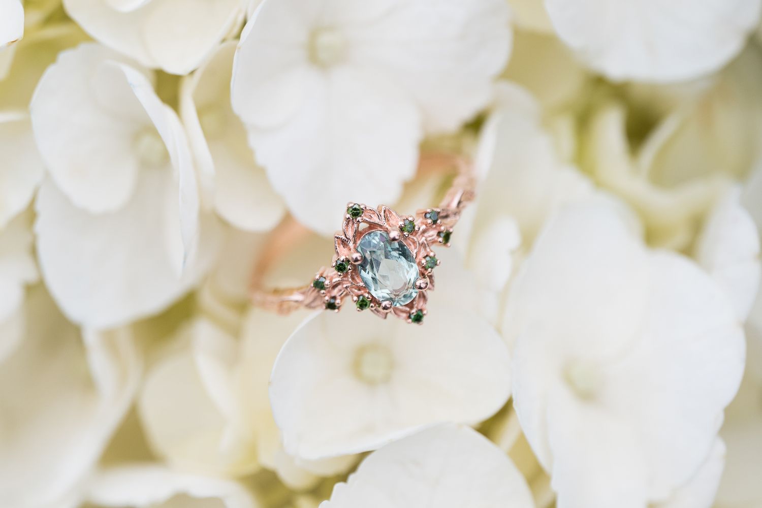 Elegant rose gold engagement ring with aquamarine center stone and floral details resting on white hydrangeas.