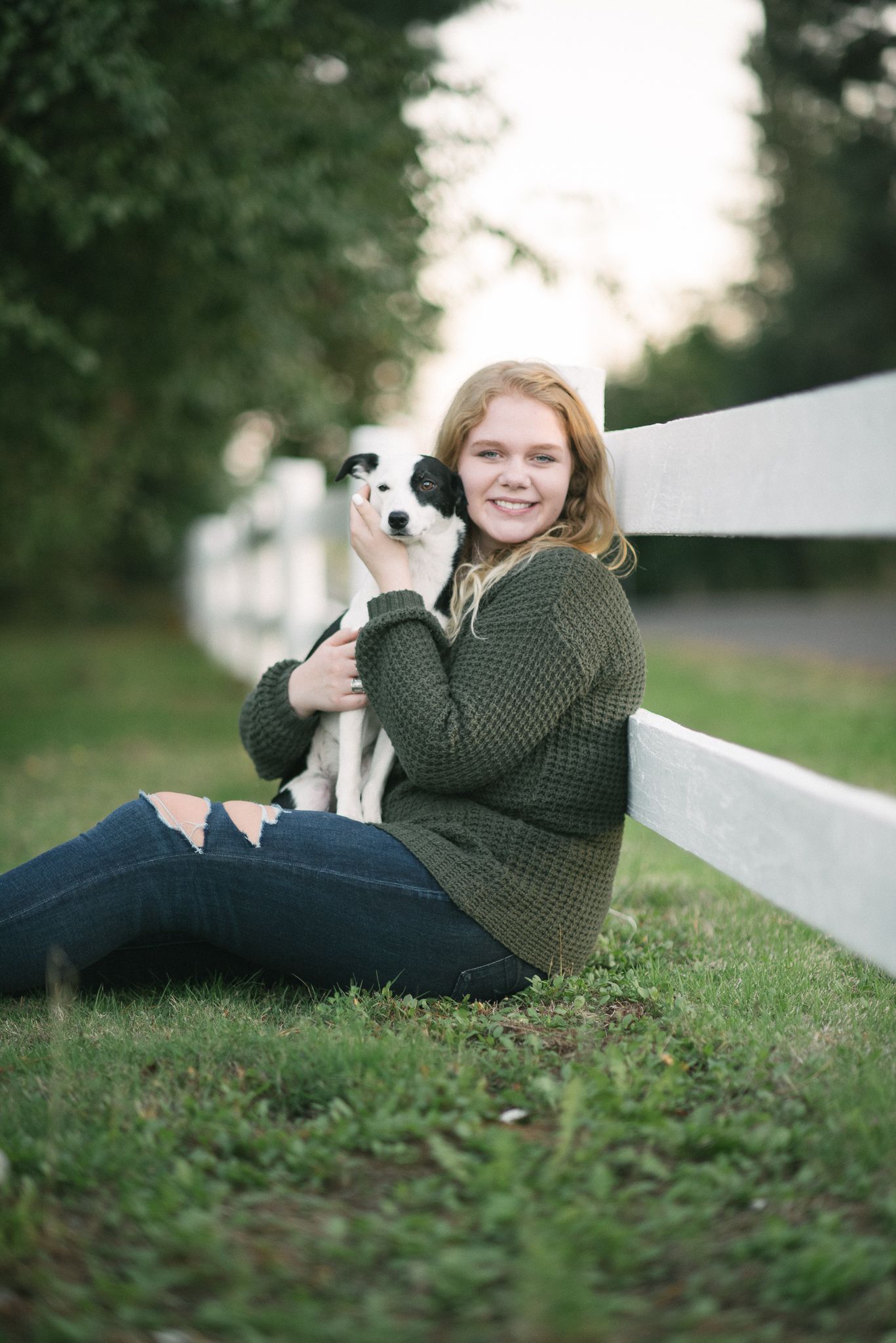 Smiling blonde teen in green sweater sitting against white fence while holding a black and white dog.