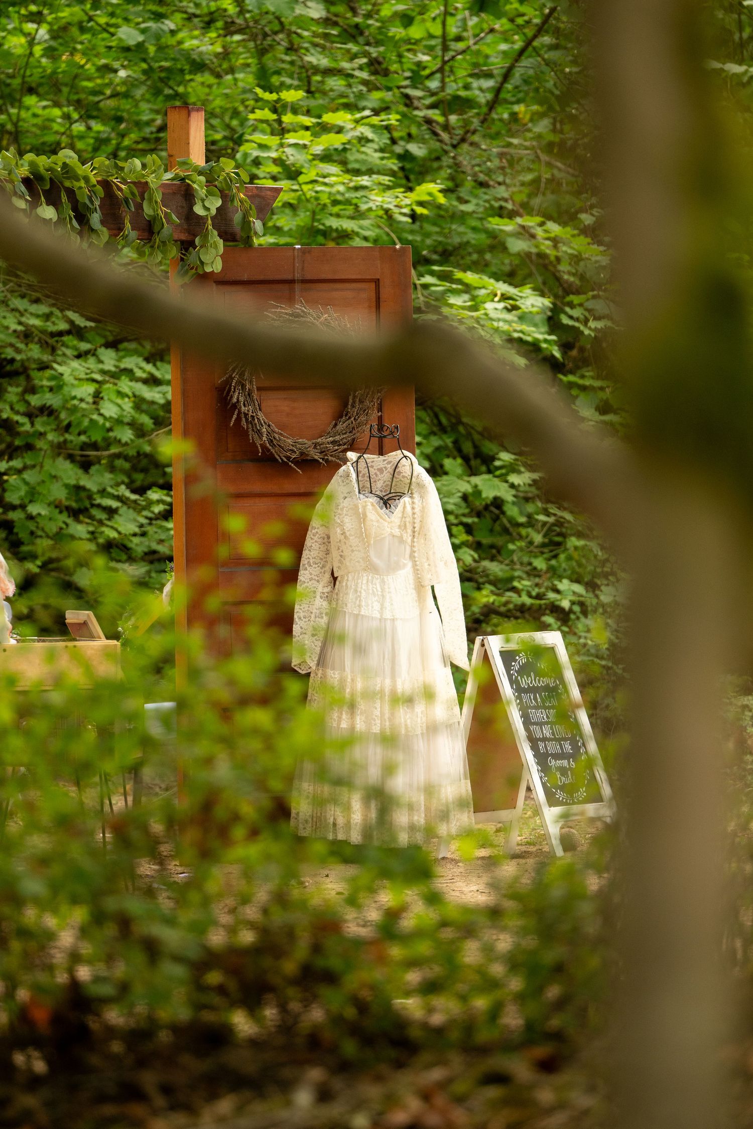 Vintage lace wedding dress displayed on rustic wooden door surrounded by lush green forest.