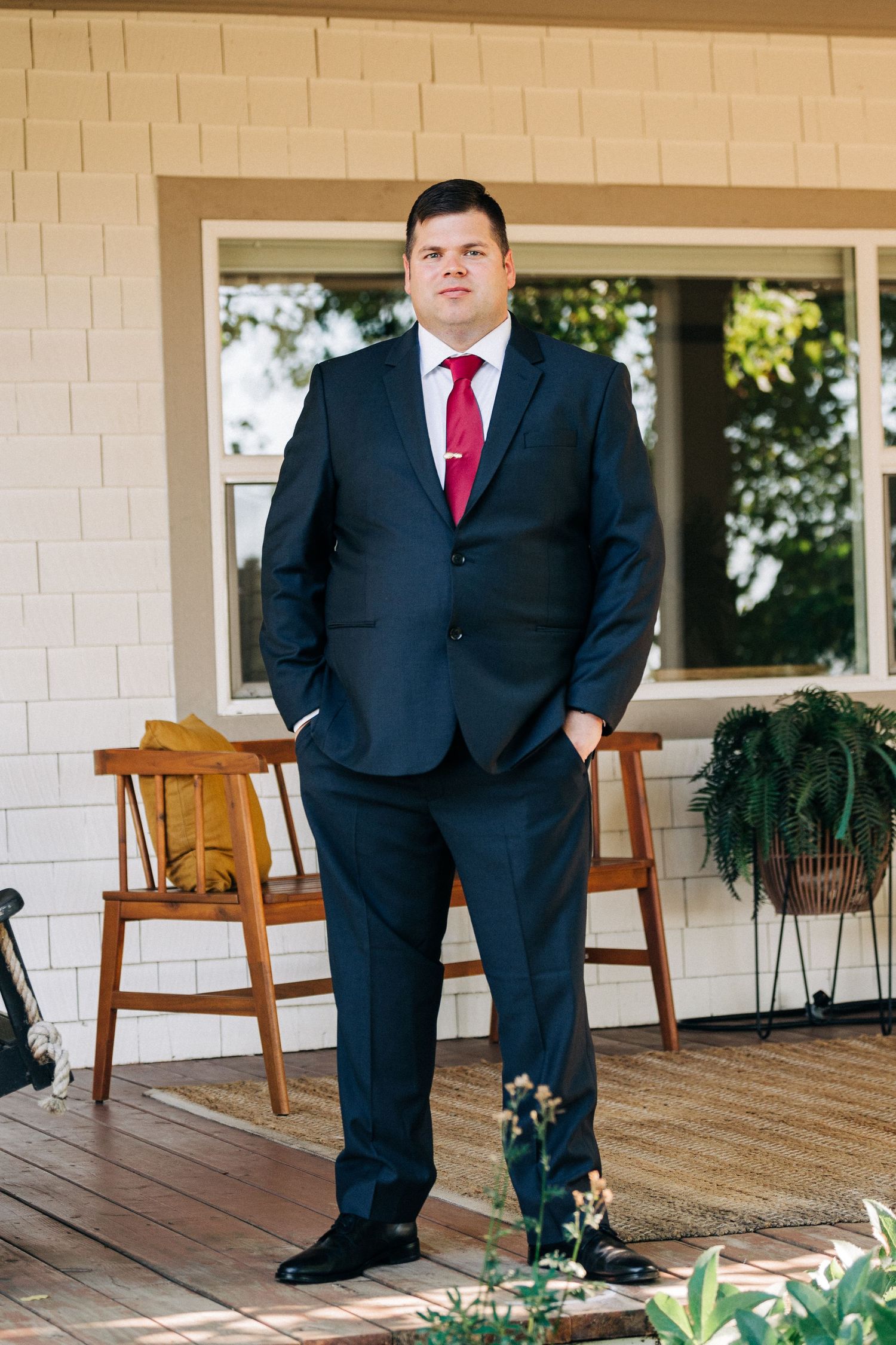 Groom in black suit and red tie poses on wooden porch with rocking chairs.