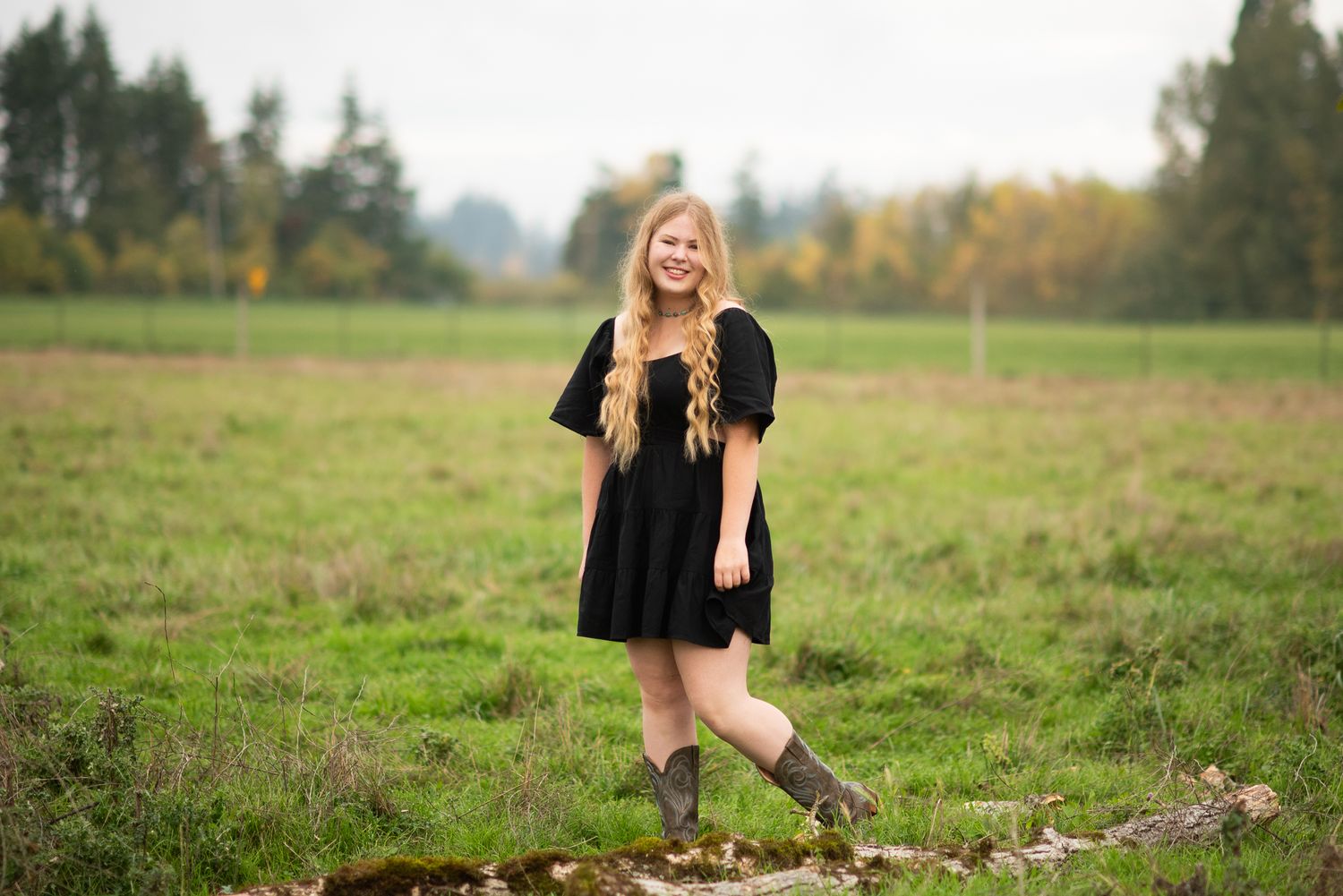 Someone wearing a black dress and boots stands in an open field during autumn.