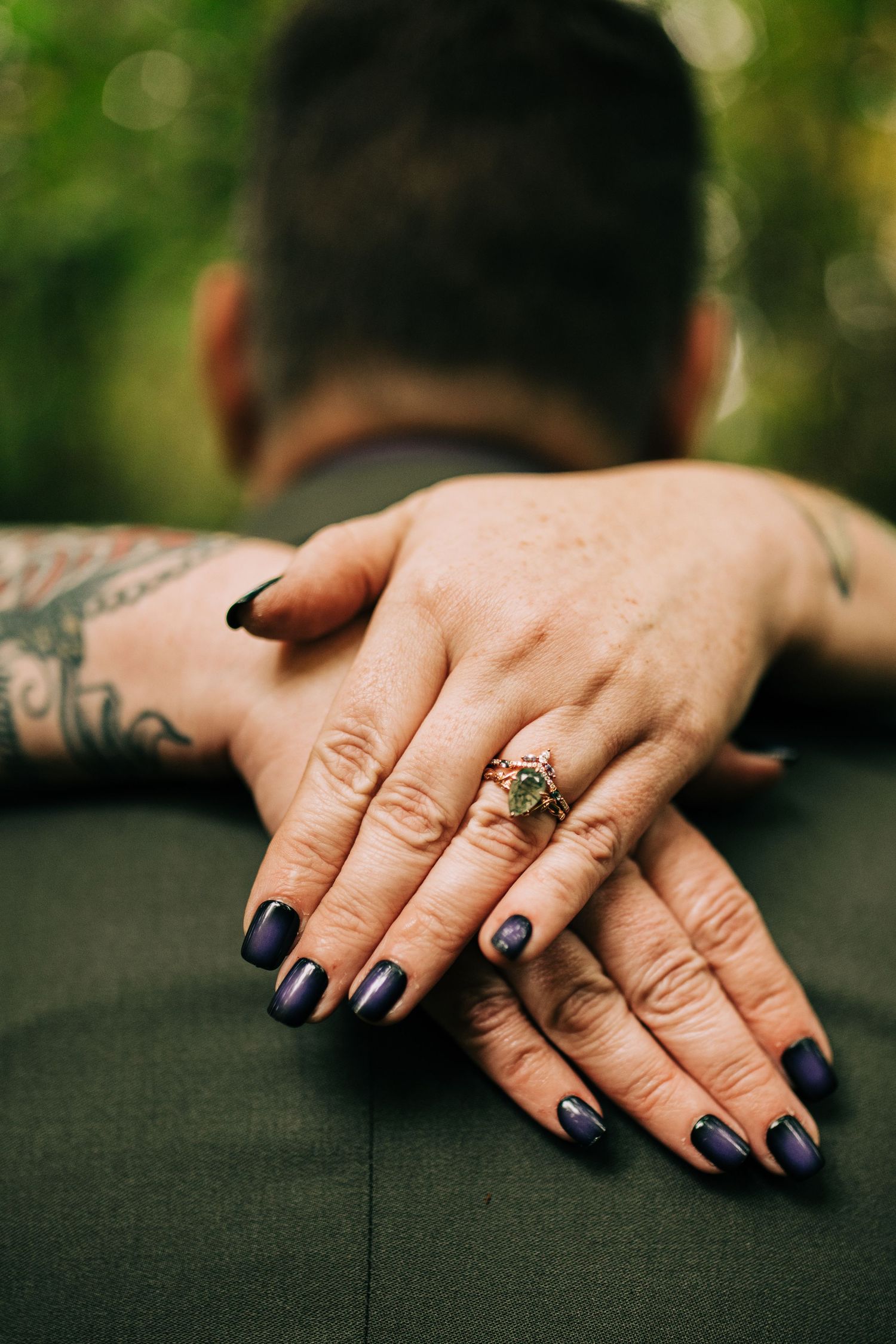 Close up of hands with dark blue nail polish and tattoos resting on a dark surface with a vintage ring.