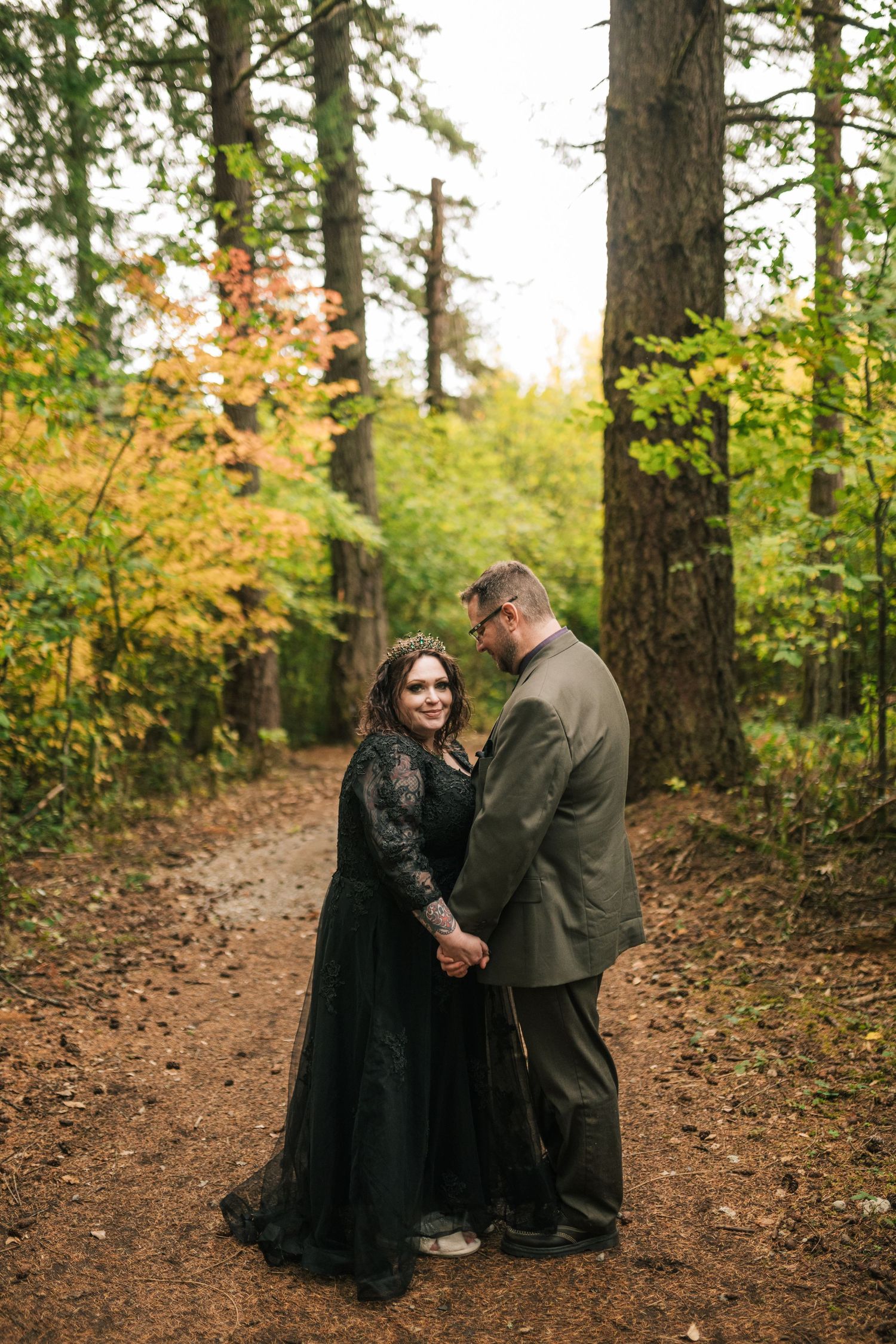 Gothic wedding couple holds hands on forest path in autumn, bride in black gown and groom in olive suit.
