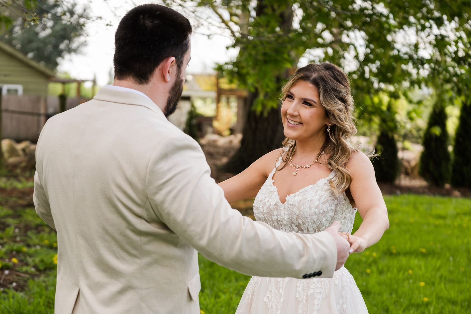 Bride in lace wedding gown smiles at groom during an outdoor first look in a lush green backyard.