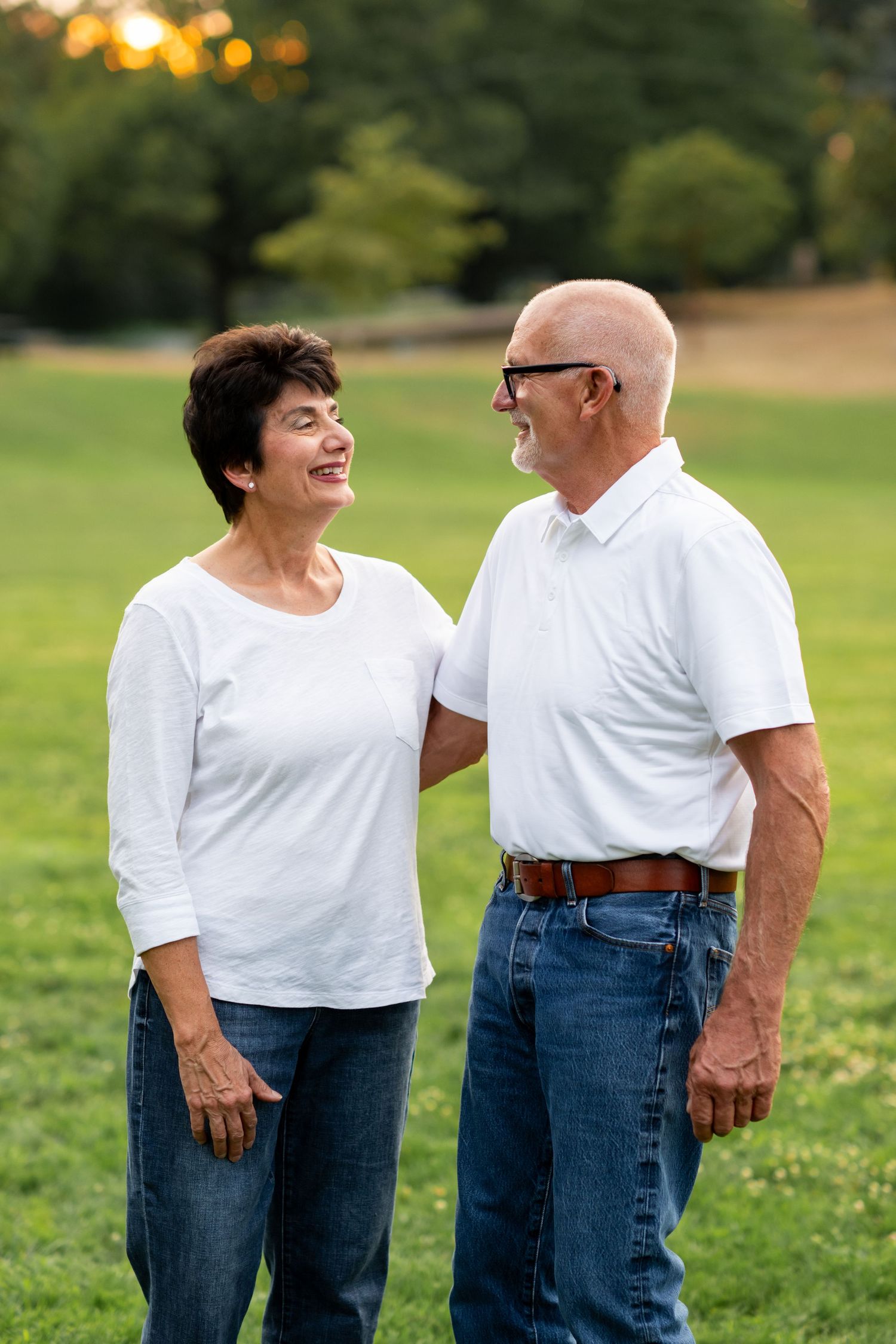 Happy senior couple in matching white shirts and jeans laughing together on a lush green lawn at sunset.