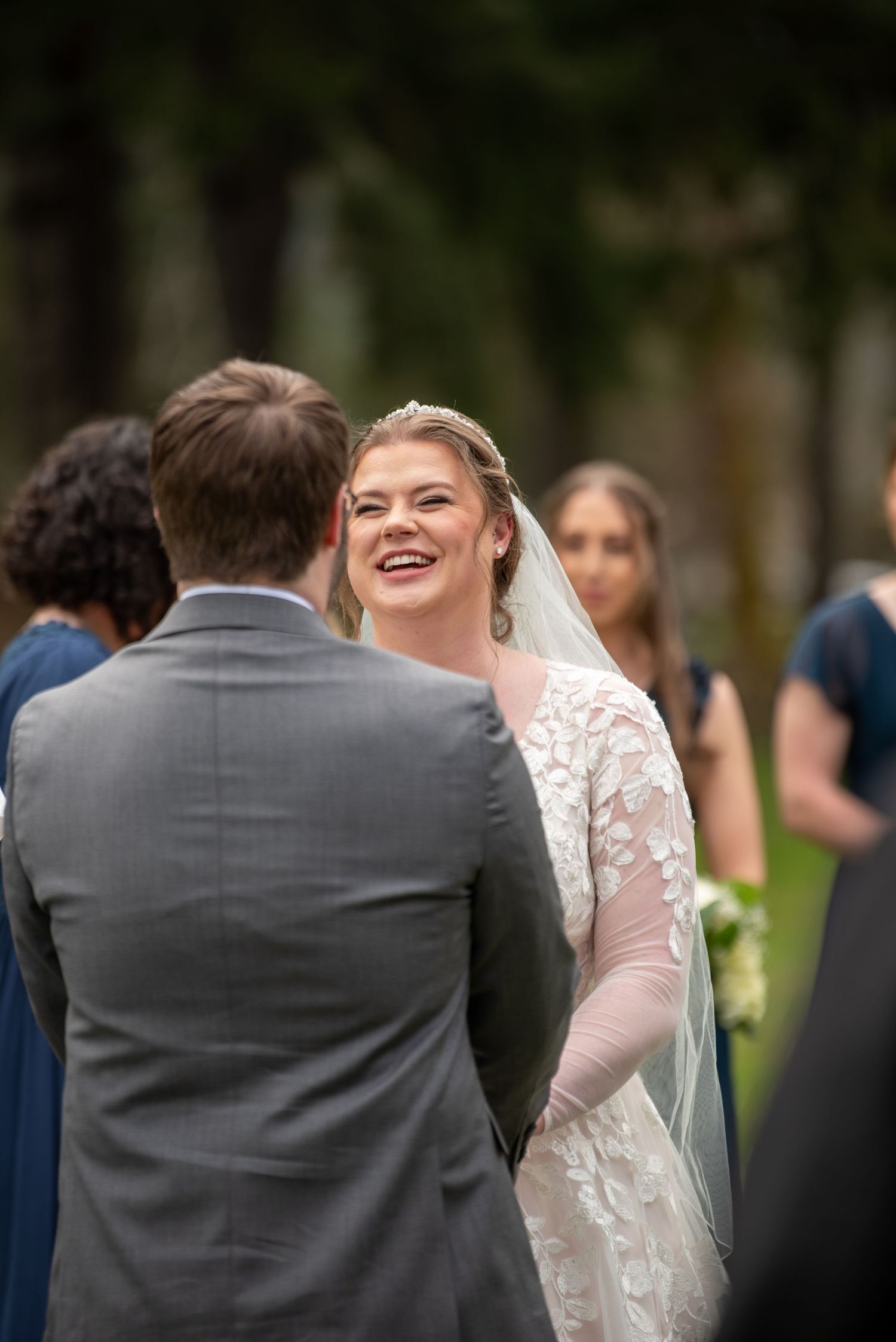 A joyful moment shared between a bride in a lace gown and groom in a grey suit during an outdoor wedding ceremony.