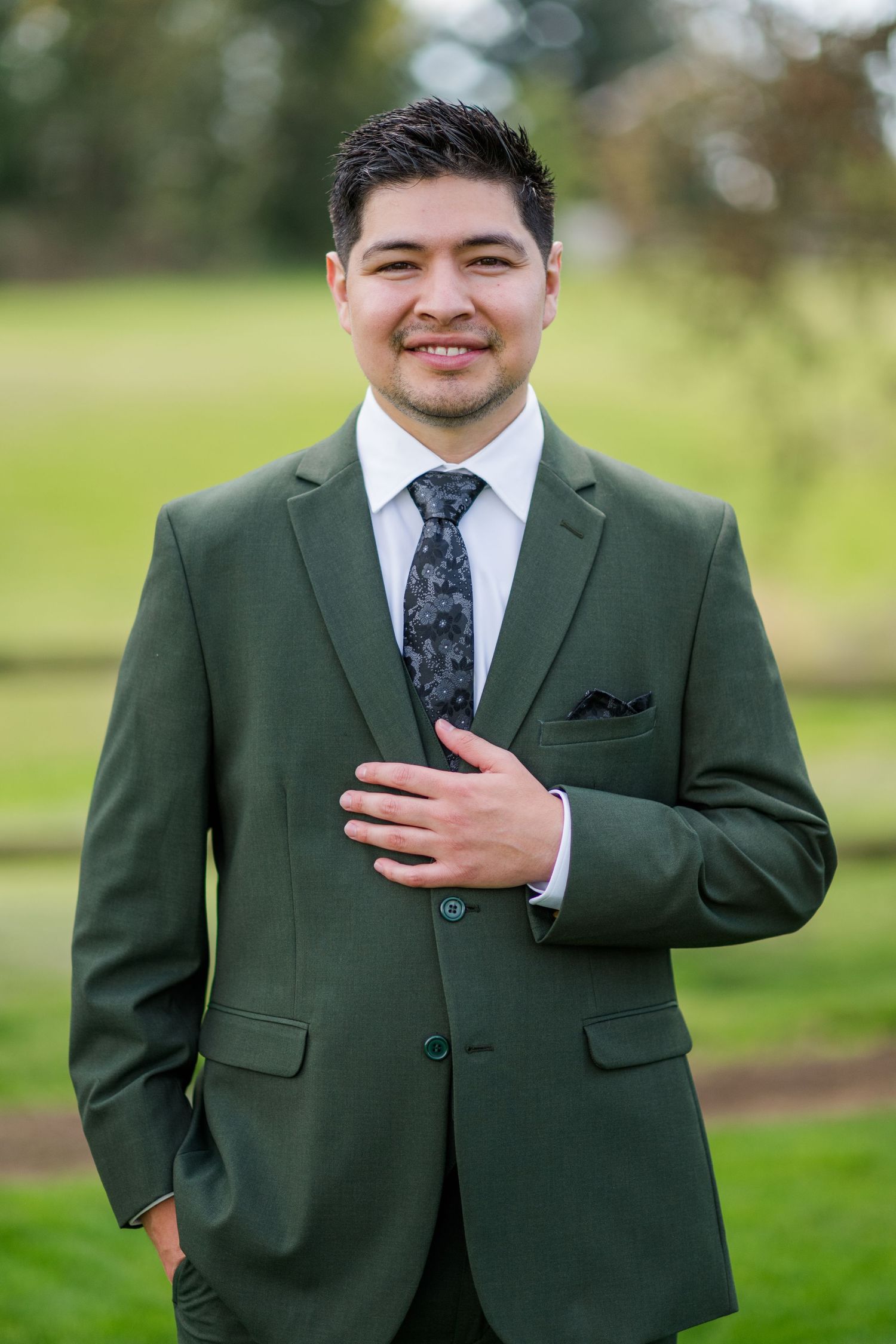 Smiling man in green suit with floral tie and pocket square posing outdoors in a park setting.