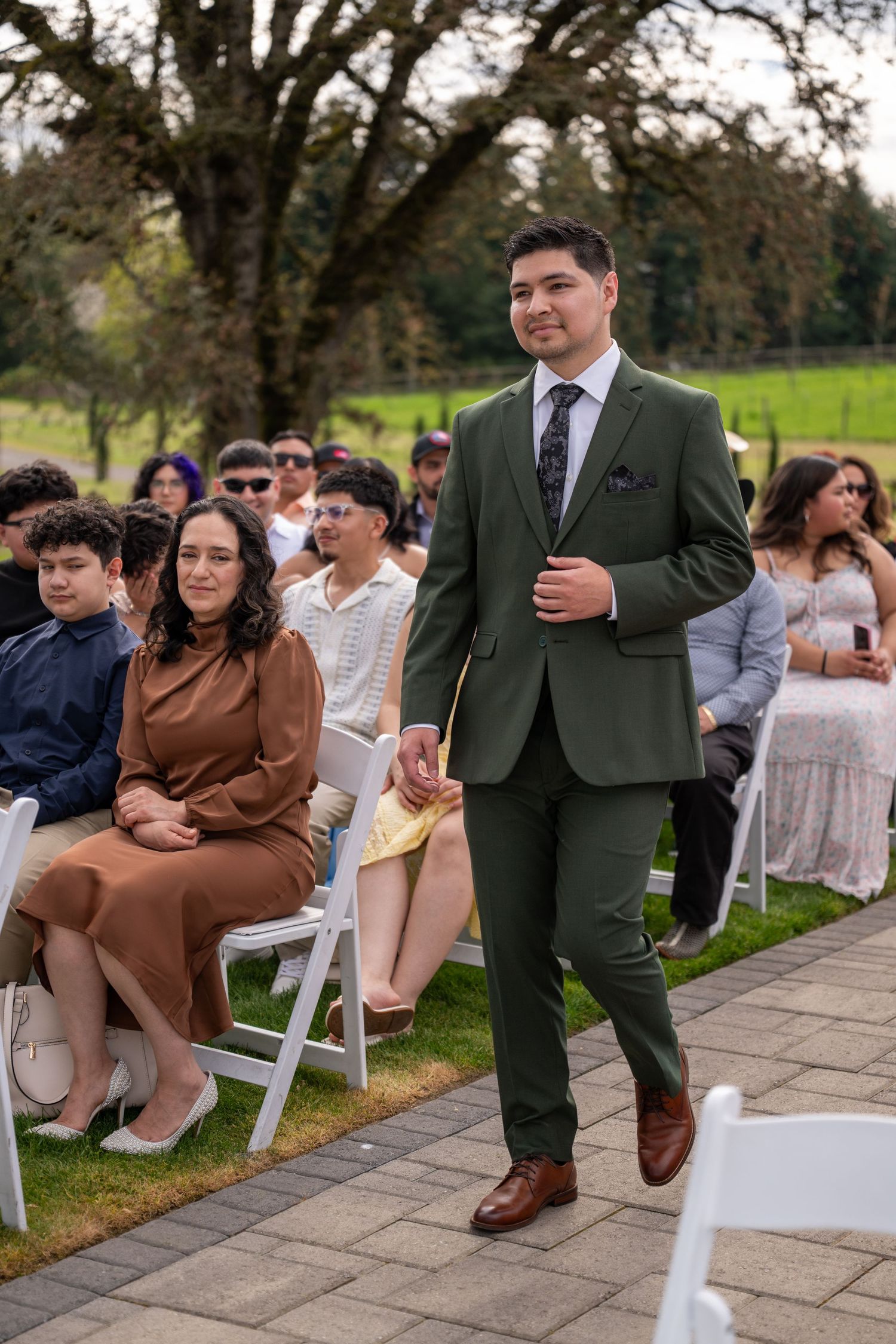 A man in a green suit walks down the aisle at an outdoor wedding ceremony with seated guests in the background.