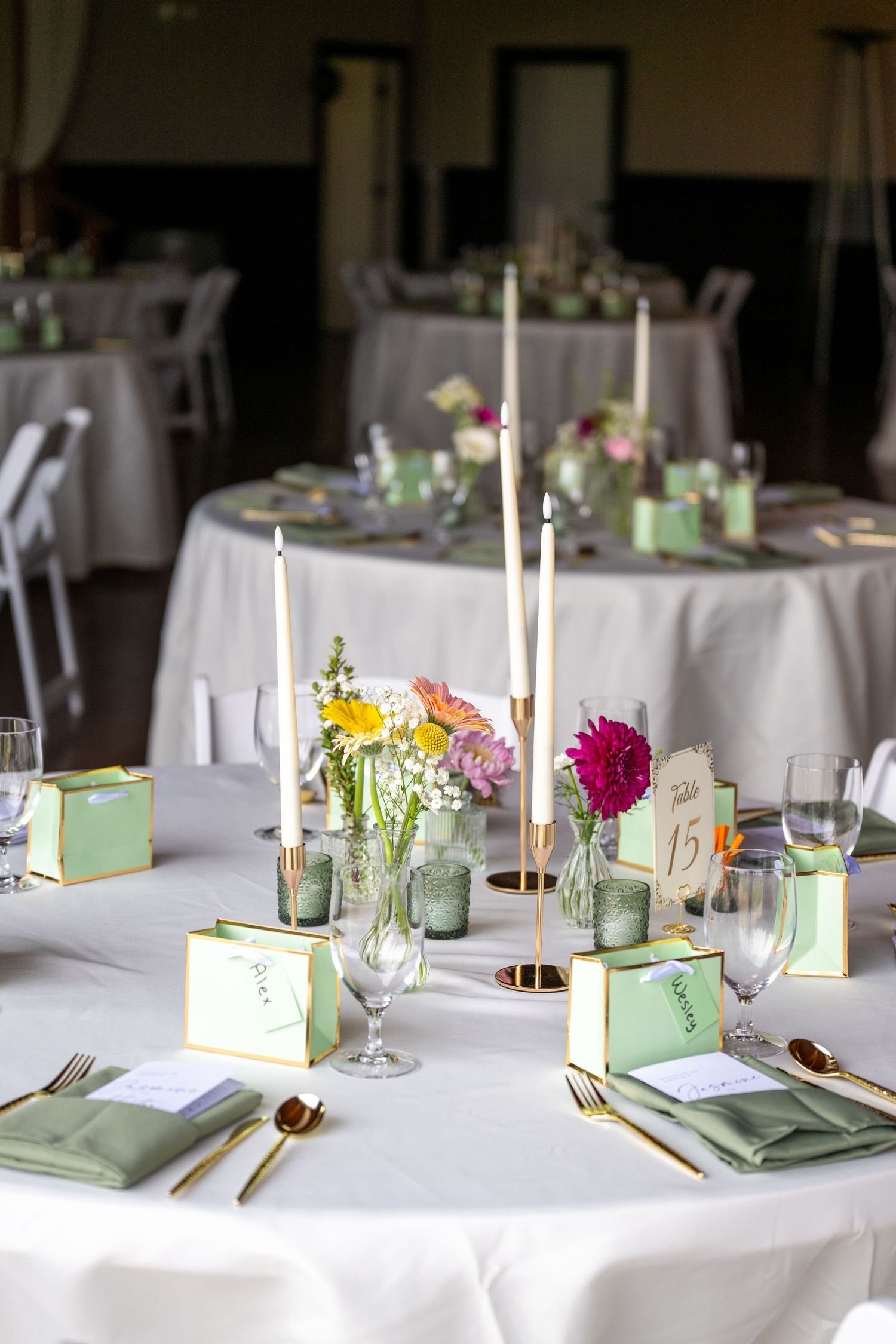 Elegant wedding reception table with white linen, gold cutlery, green napkins, candles, and colorful floral centerpieces.