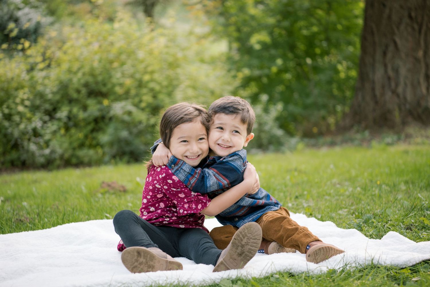Two young siblings sitting on a white blanket in a park, smiling and hugging each other warmly.