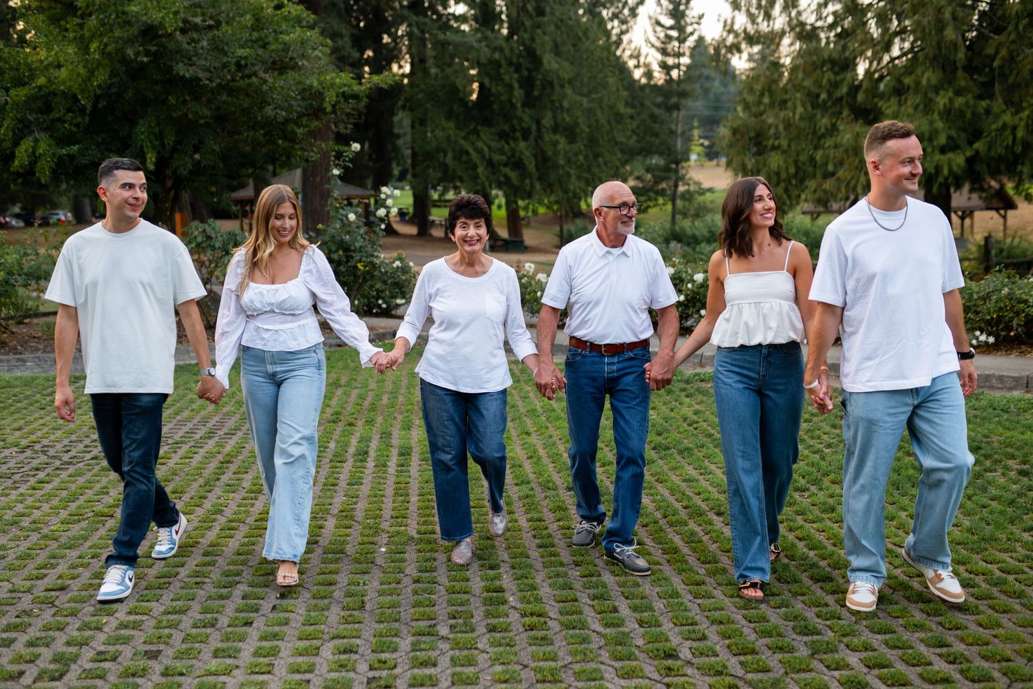Family of six holding hands and walking together in white tops and jeans in a park.