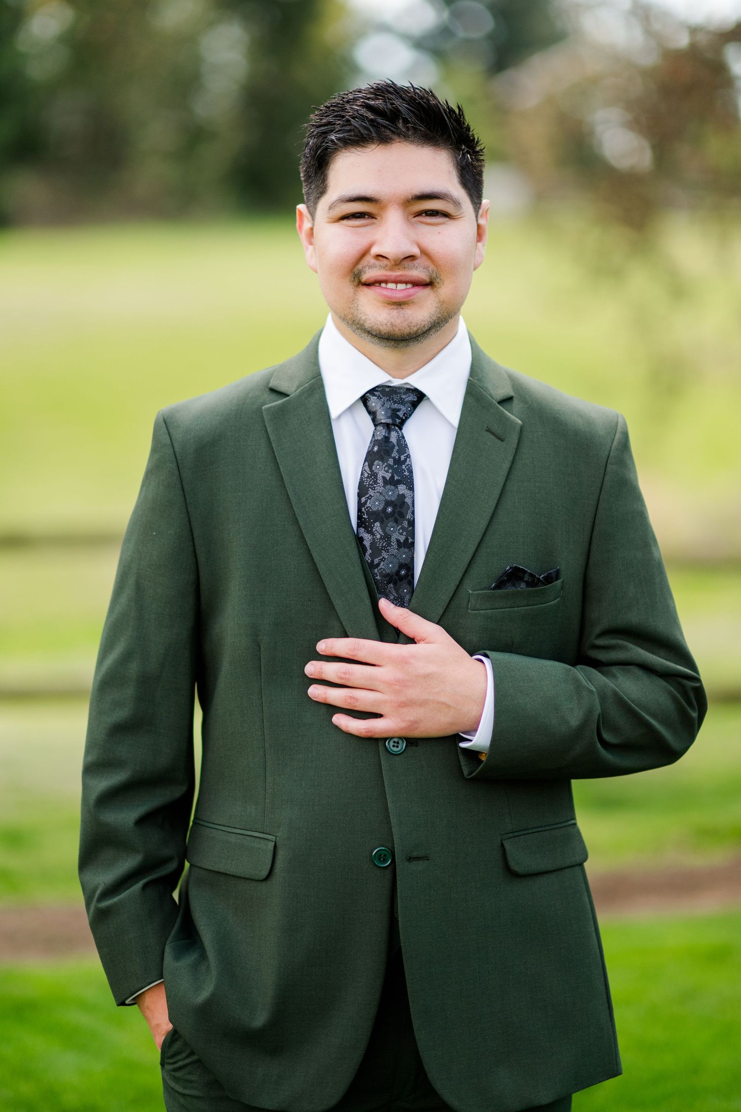 Smiling groomsman in olive green suit with dark floral tie poses confidently outdoors at a wedding.