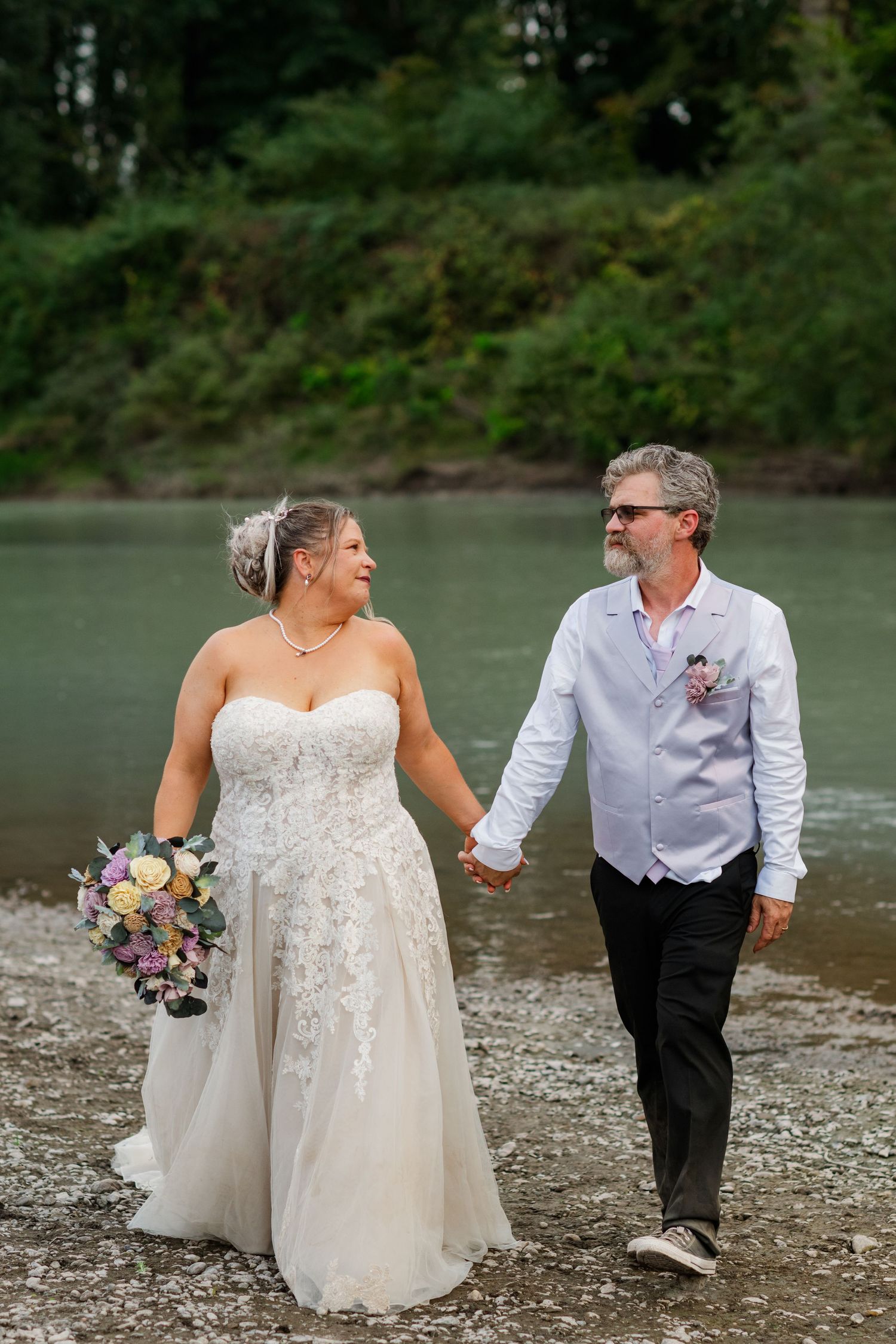 Bride in lace wedding gown and groom in white shirt walk hand-in-hand along scenic riverside wedding venue.
