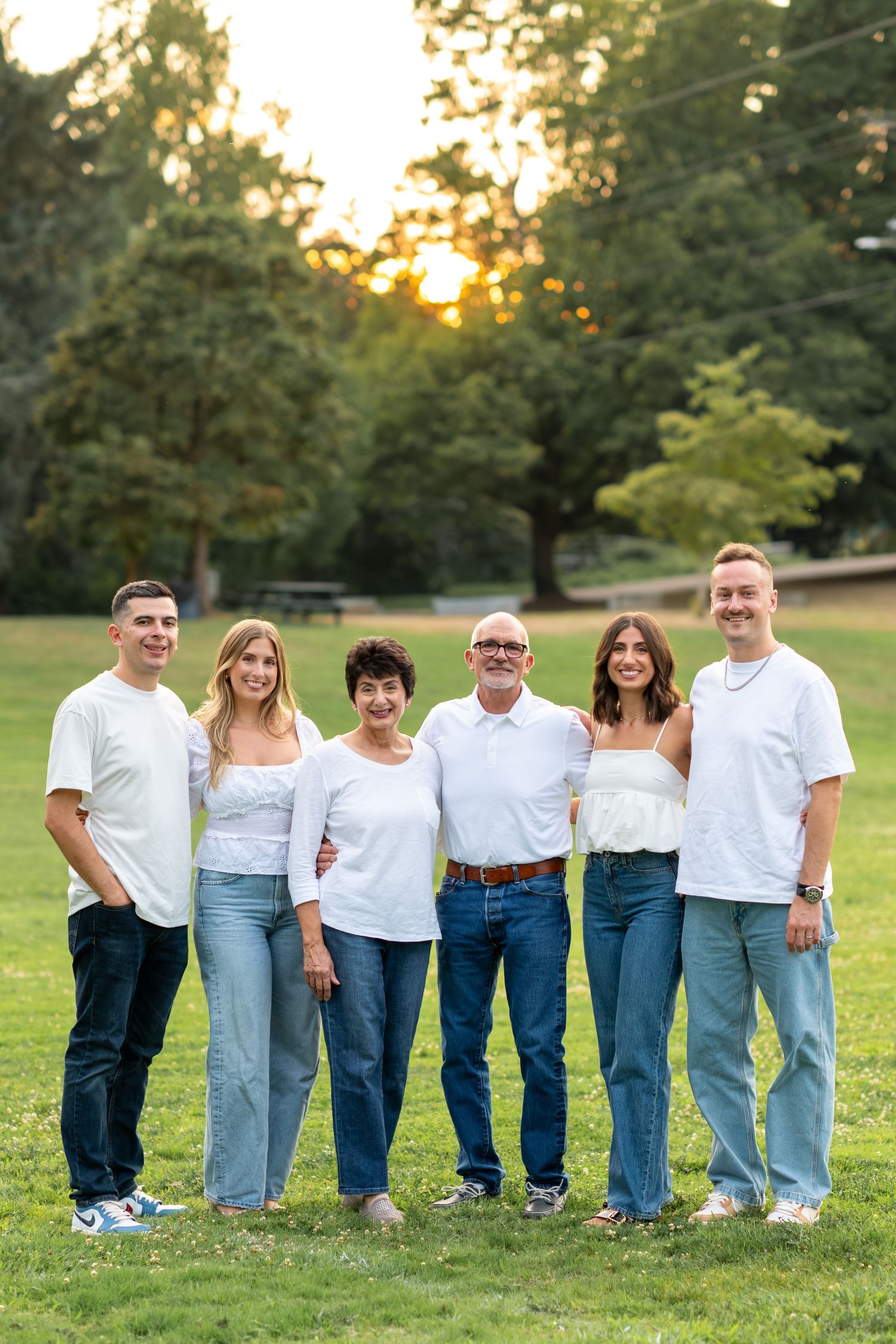 A family of six in matching white tops and jeans poses together at sunset in a park.