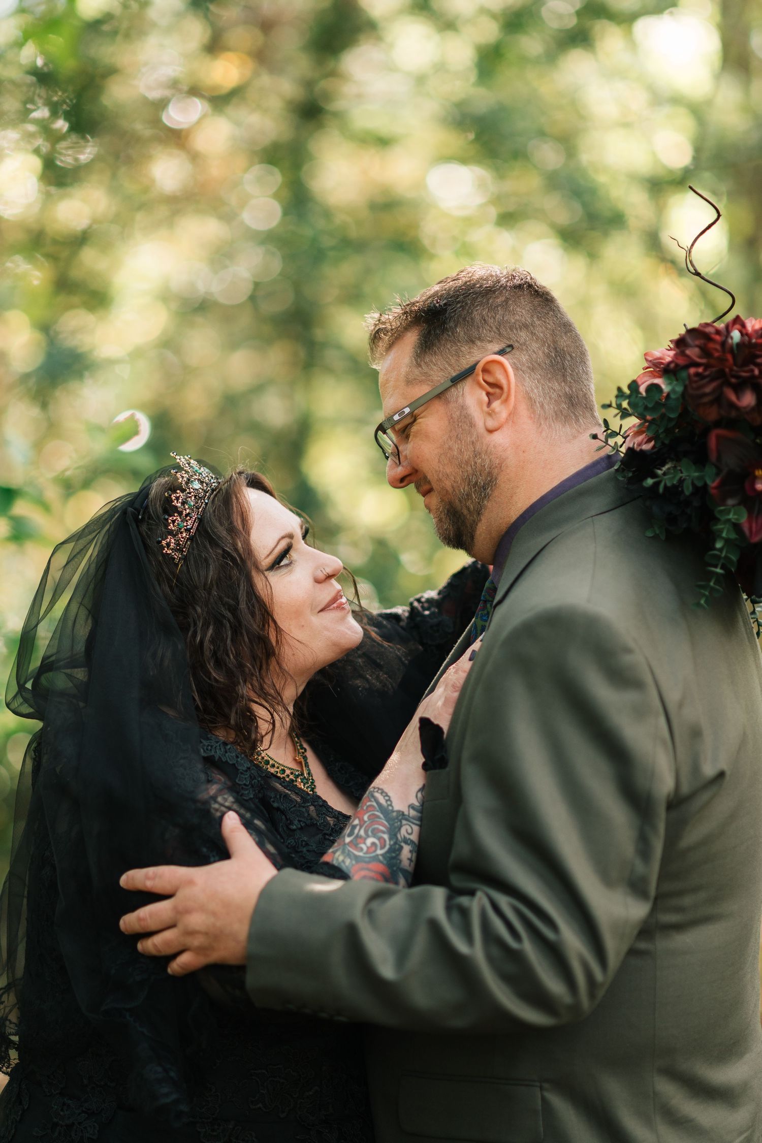 Gothic wedding couple embracing in sunlit forest, bride in black lace dress with jeweled crown smiles at groom.