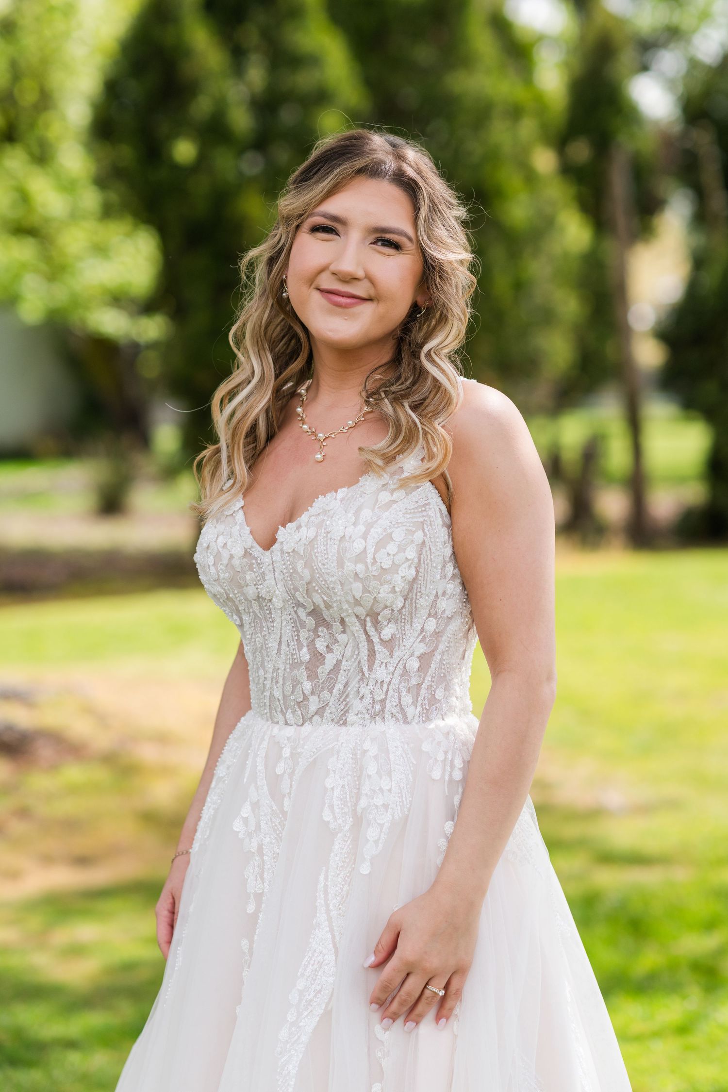 Bride in strapless lace wedding gown smiles softly outdoors surrounded by lush green trees.