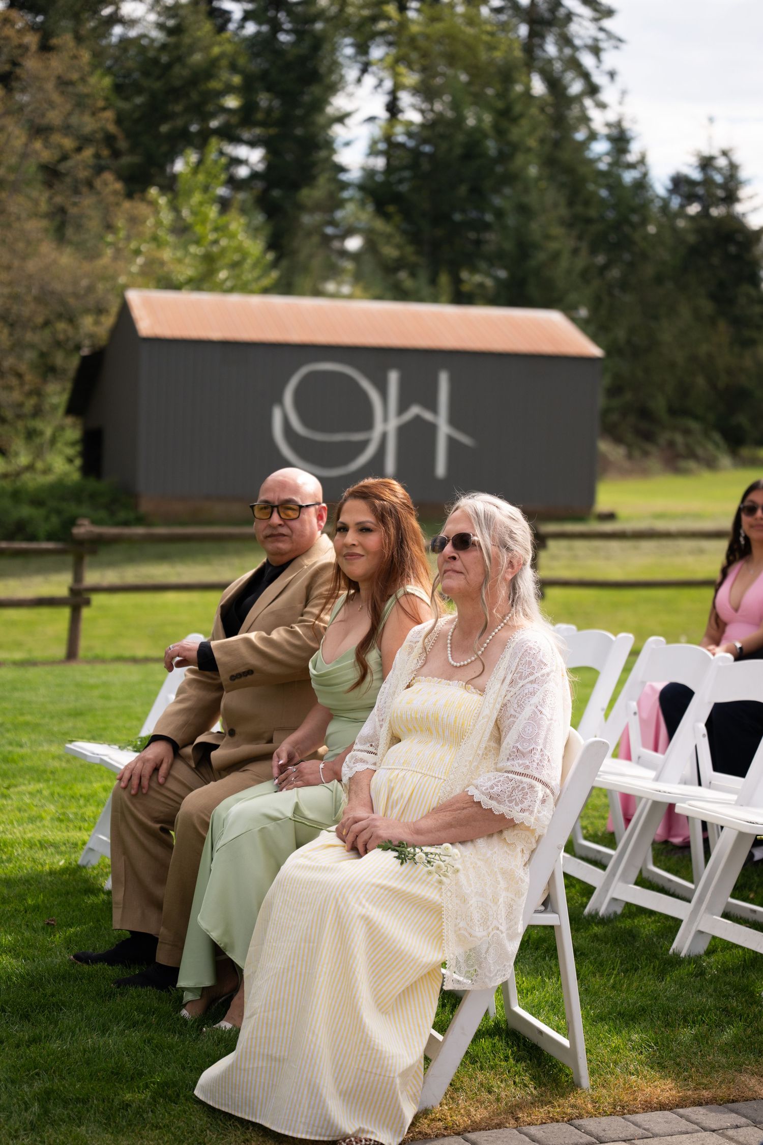 Guests seated in white chairs at an outdoor wedding ceremony on a sunny day with a barn and trees in the background.