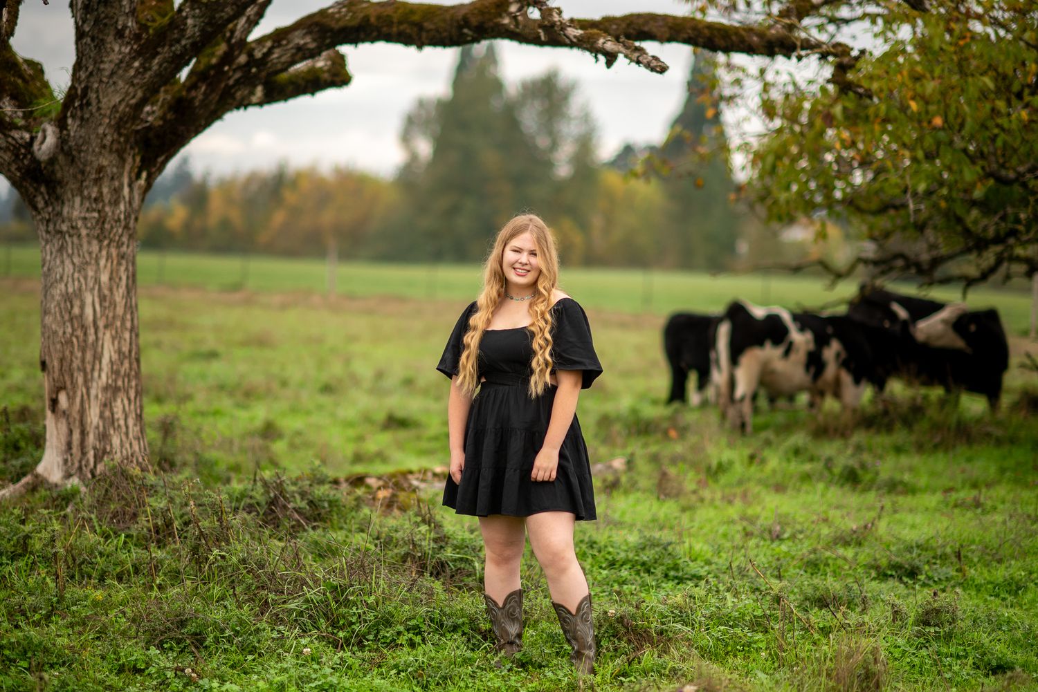 Smiling blonde teen in black dress and cowboy boots stands under a tree with cattle grazing nearby.