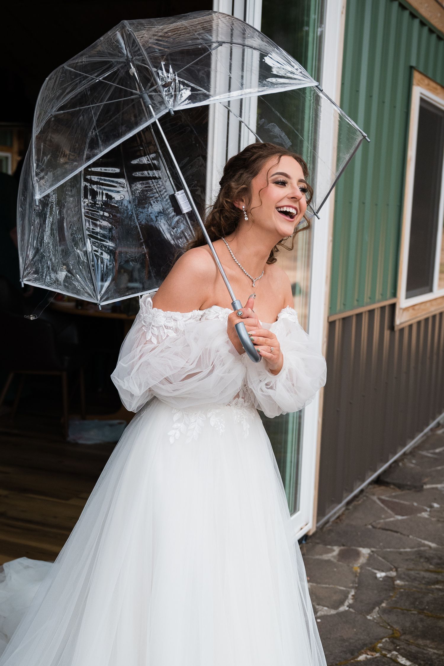 Laughing bride in white off-shoulder gown holds clear umbrella outside green building on rainy day.