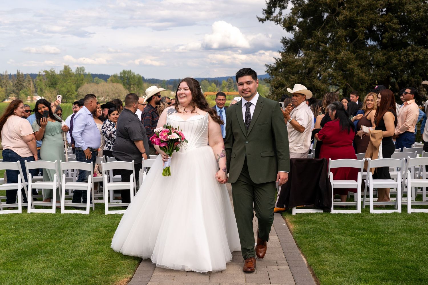 Bride and groom walk hand-in-hand down the aisle after outdoor wedding ceremony with guests seated on white chairs.