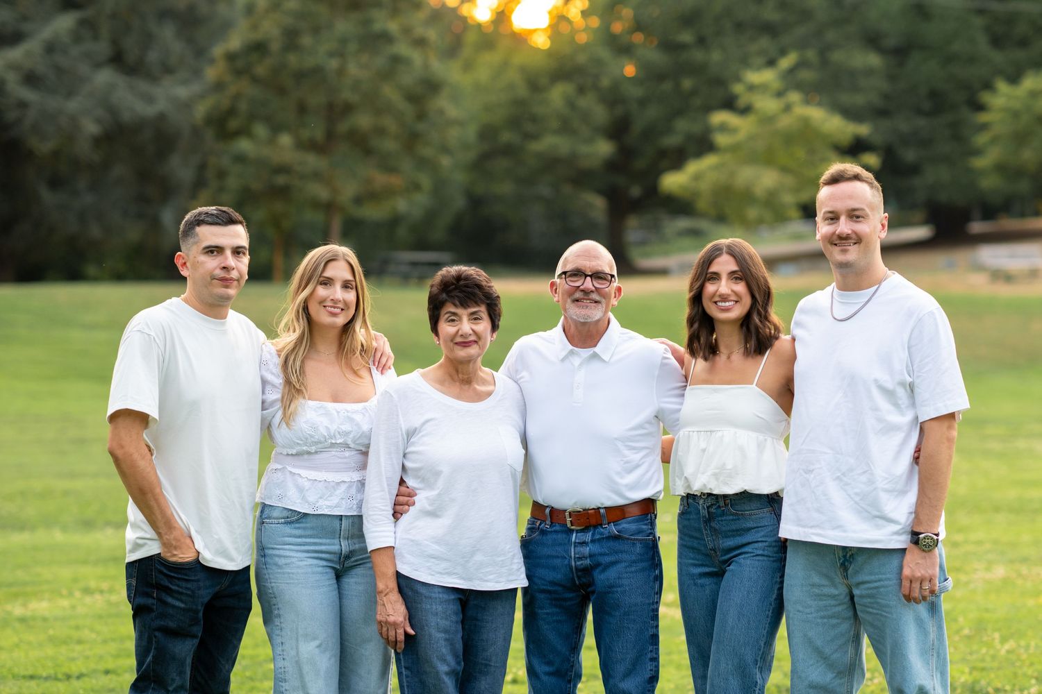 Family of six smiling outdoors in white tops and jeans in a green park setting.
