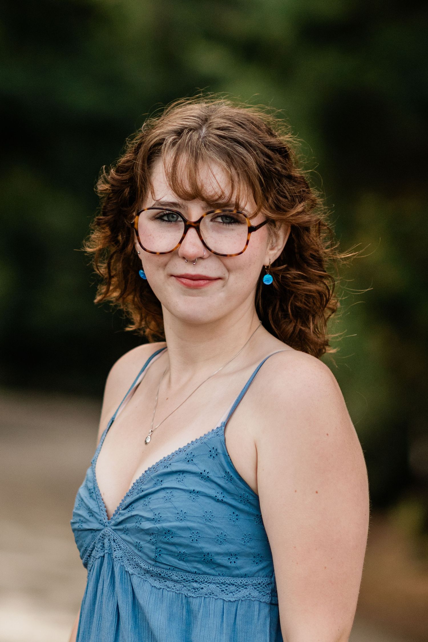 Young woman with curly auburn hair and glasses wearing a blue sundress posing outdoors in natural light.