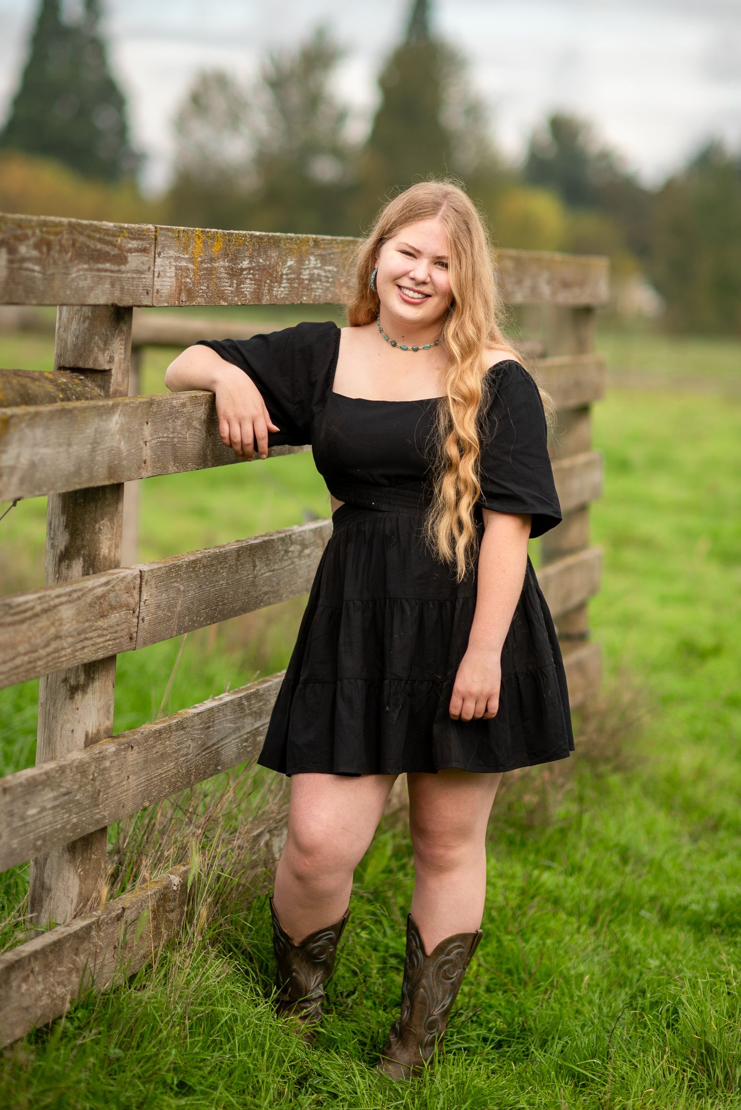 Smiling blonde teen in black dress and cowboy boots leans against a rustic wooden farm fence.