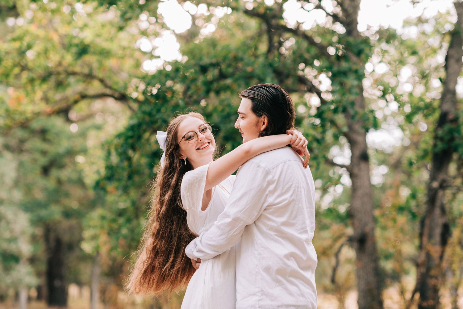 Couple in matching white outfits embrace and smile at each other in a sunlit forest setting.
