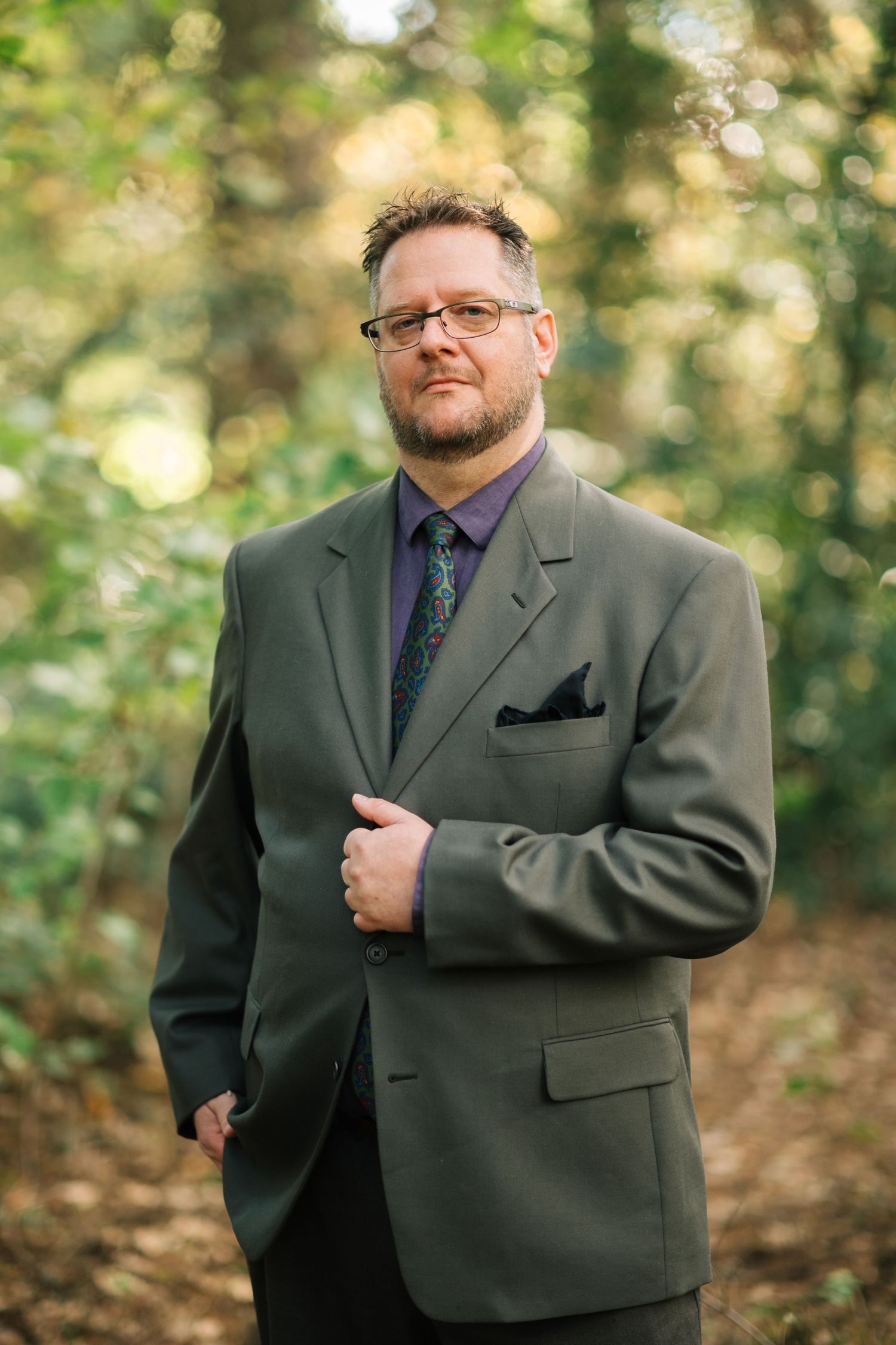A person wearing a green suit jacket and tie poses outdoors in a wooded setting with natural bokeh lighting effects.