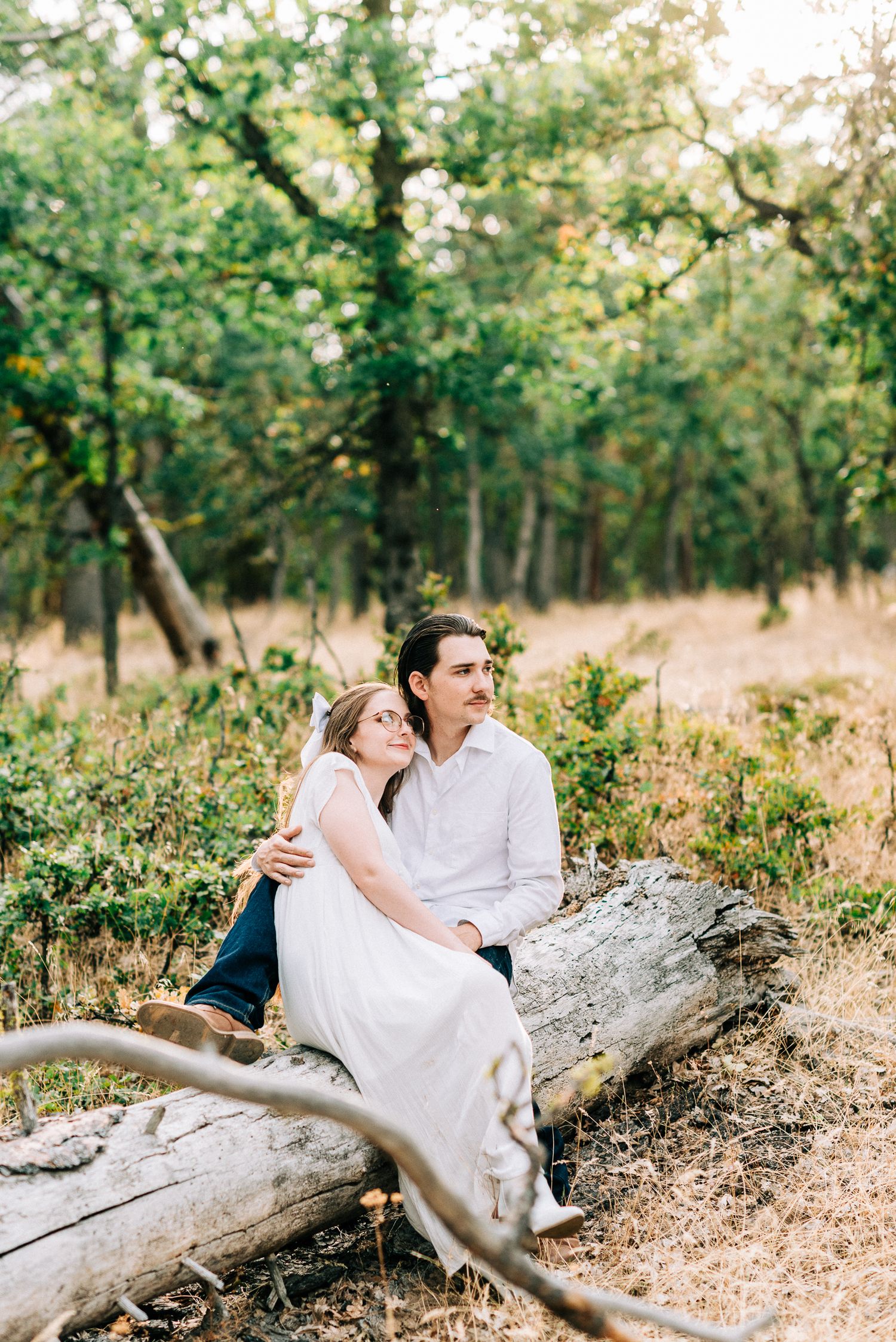 A bride and groom sit together on a fallen log in a peaceful woodland setting.