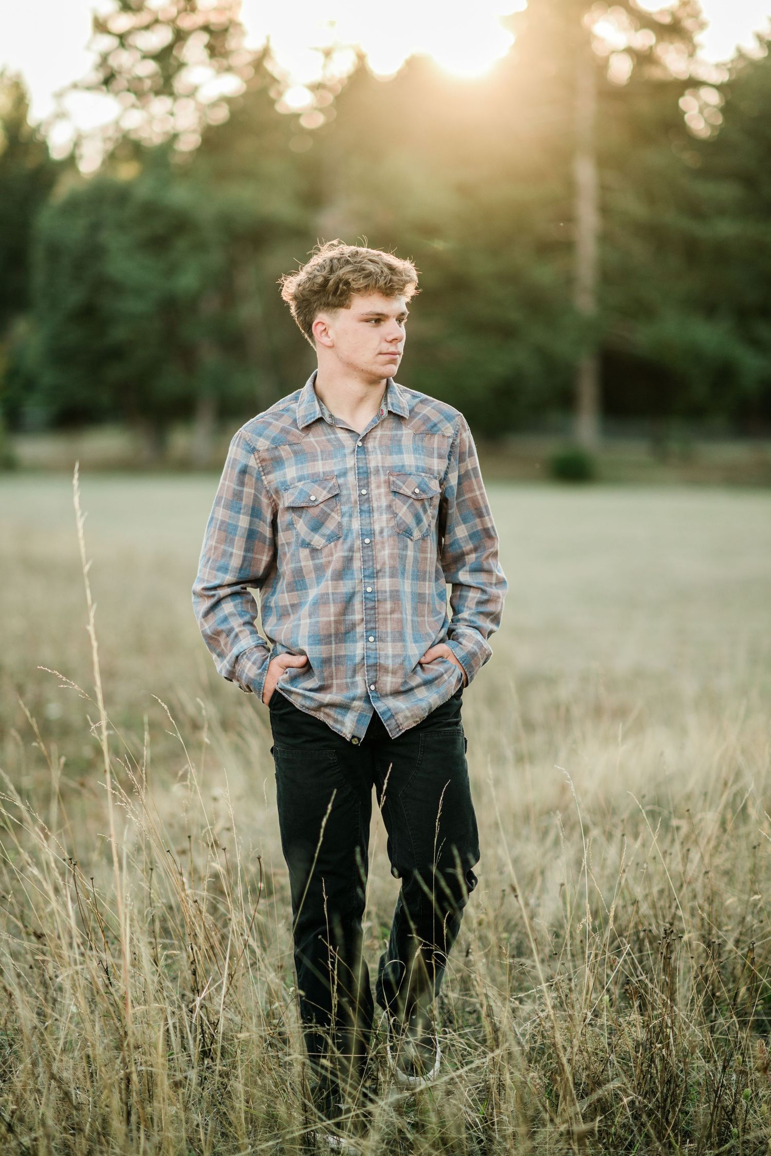 Teenage boy in a plaid shirt standing in tall grass with hands in pockets during a golden sunset outdoors.