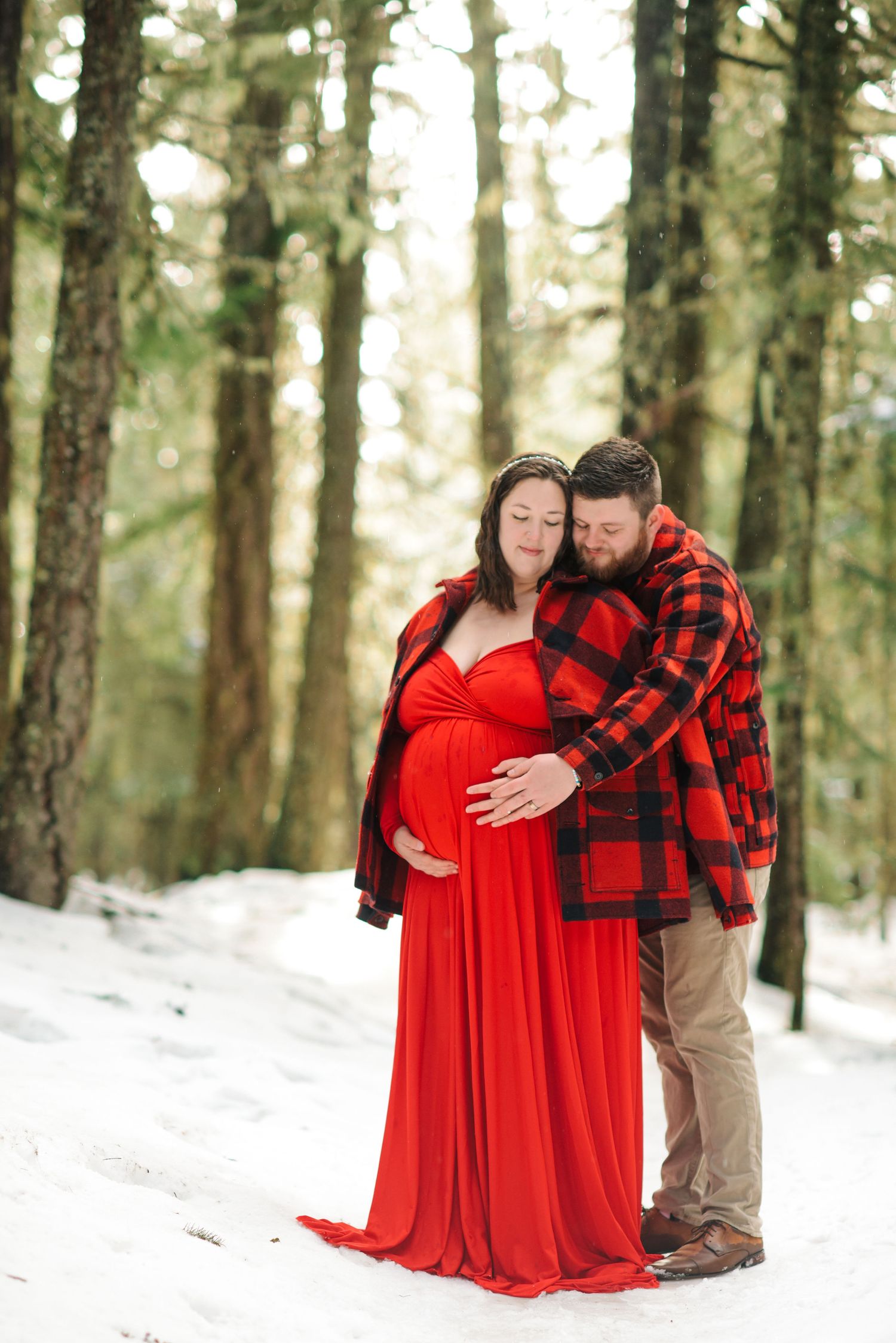 Intimate embrace captured in winter forest with red dress and plaid shirt against snow.