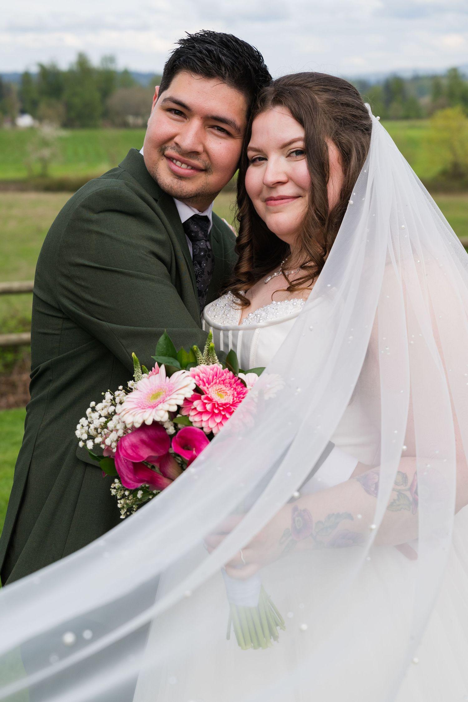 Smiling bride and groom pose outdoors, bride holding pink bouquet with veil flowing, groom in green suit.
