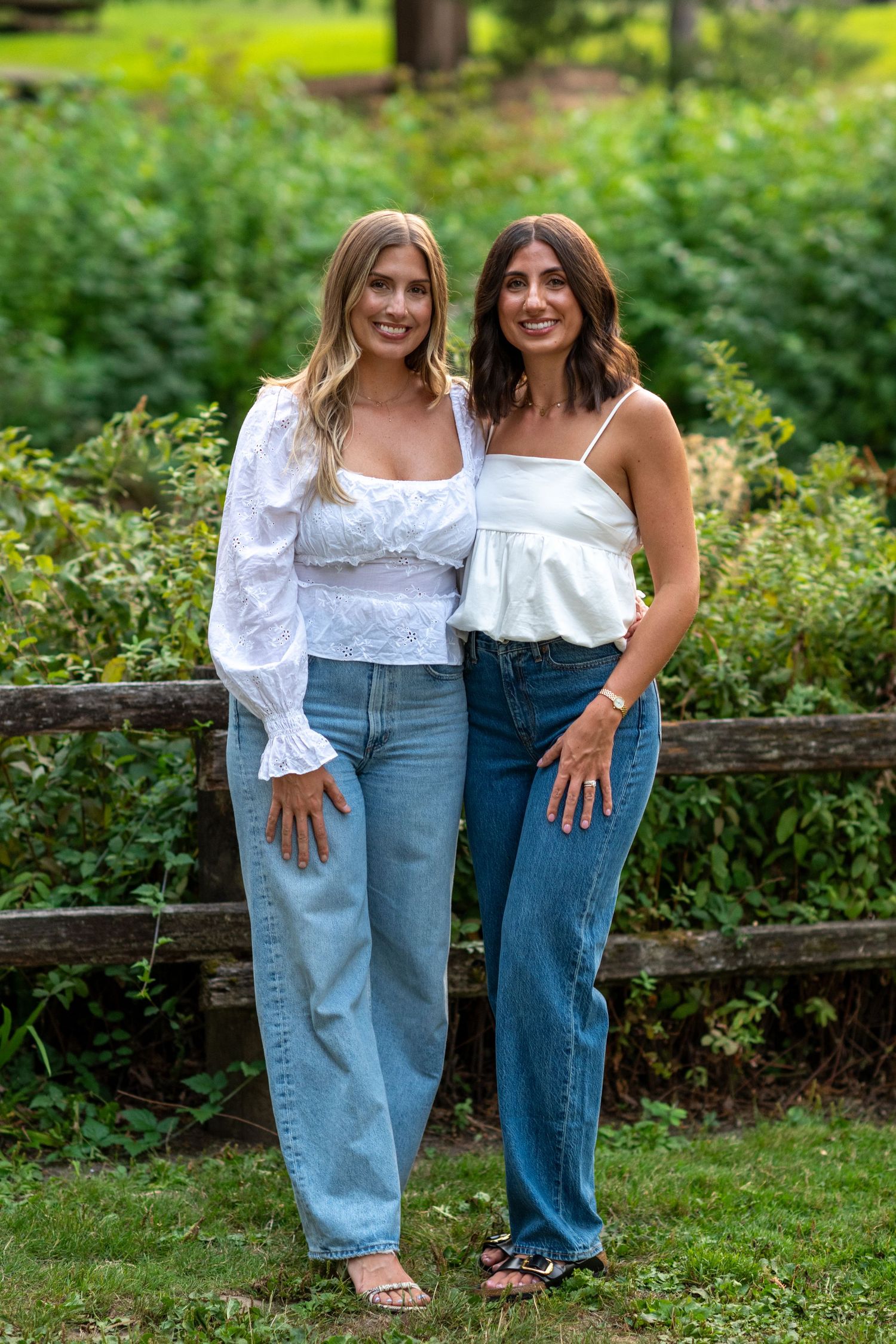 Two young women in white tops and jeans smiling together by a wooden fence outdoors.