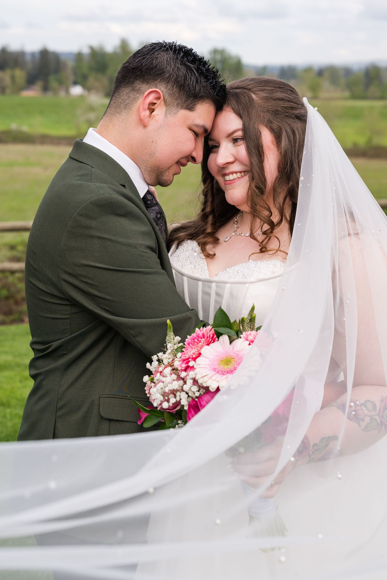 Bride smiling joyfully as groom touches foreheads with her, veil flowing in the breeze.