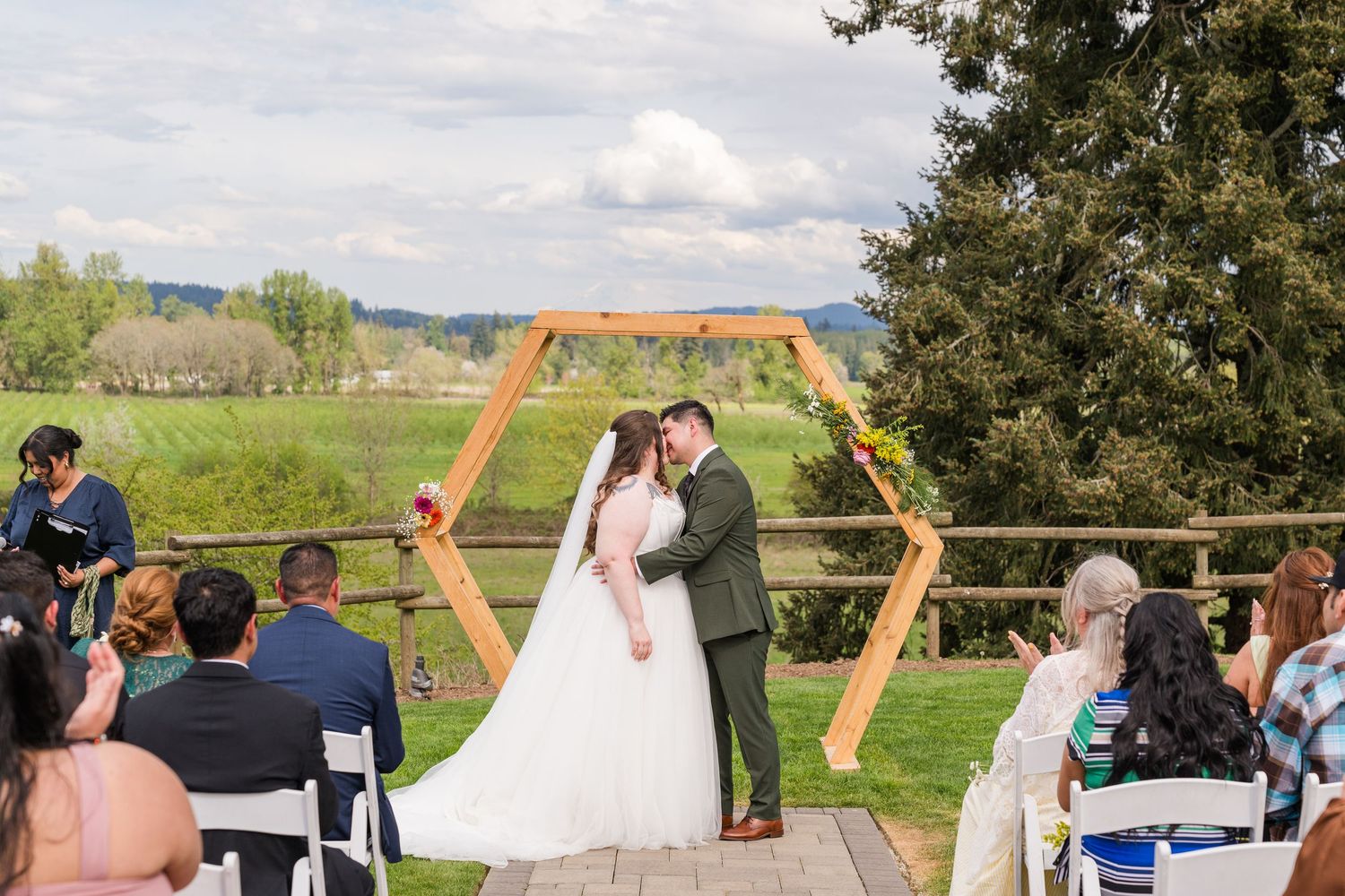 Bride and groom share first kiss at outdoor ceremony beneath hexagonal wooden arch with scenic green valley view.