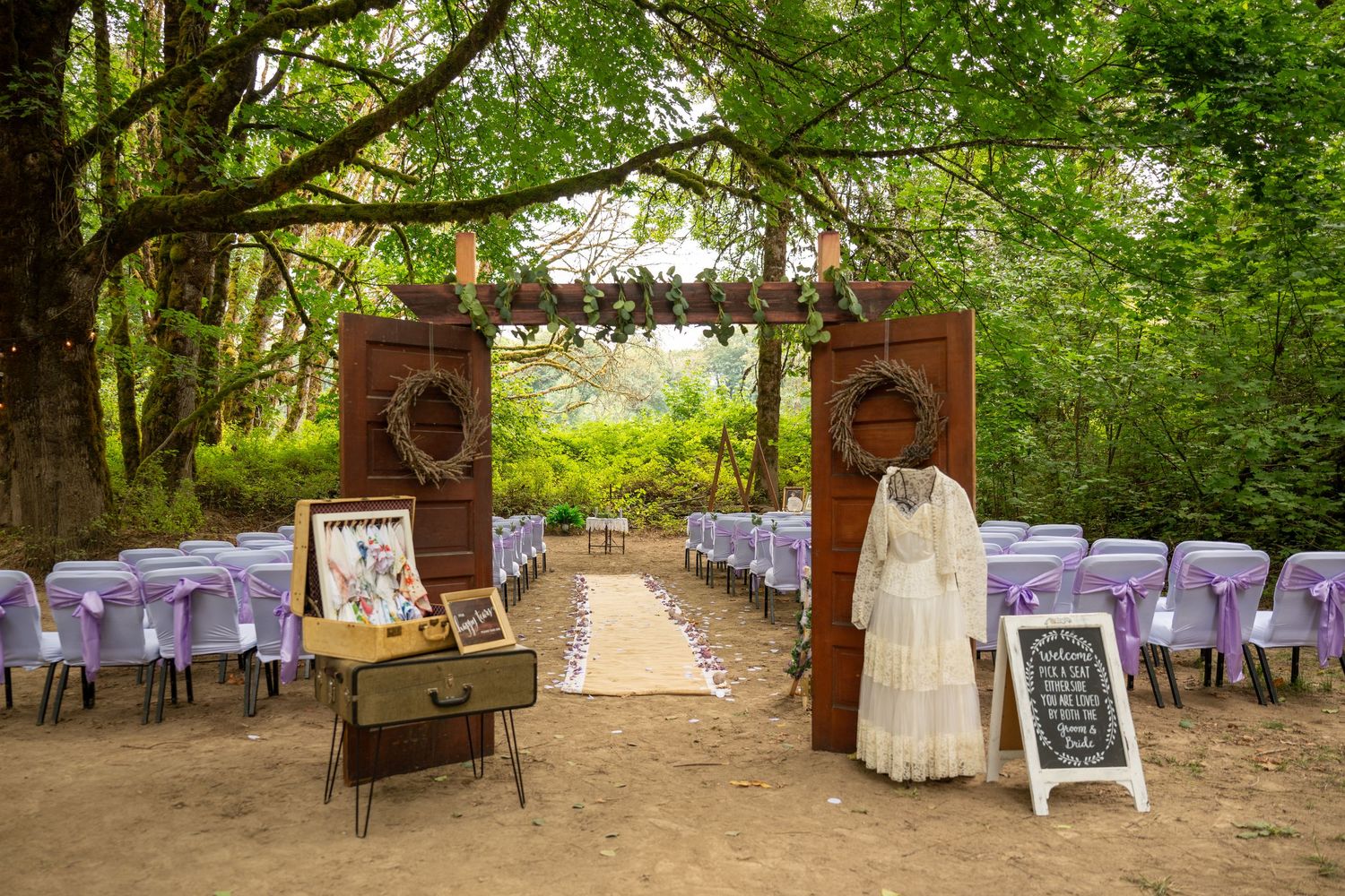 Rustic outdoor wedding ceremony setup with wooden arch, purple chairs, and white dress display.