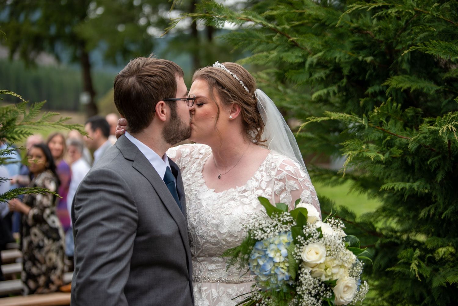 Newlyweds sharing their first kiss as a married couple outdoors surrounded by lush greenery and wedding guests.