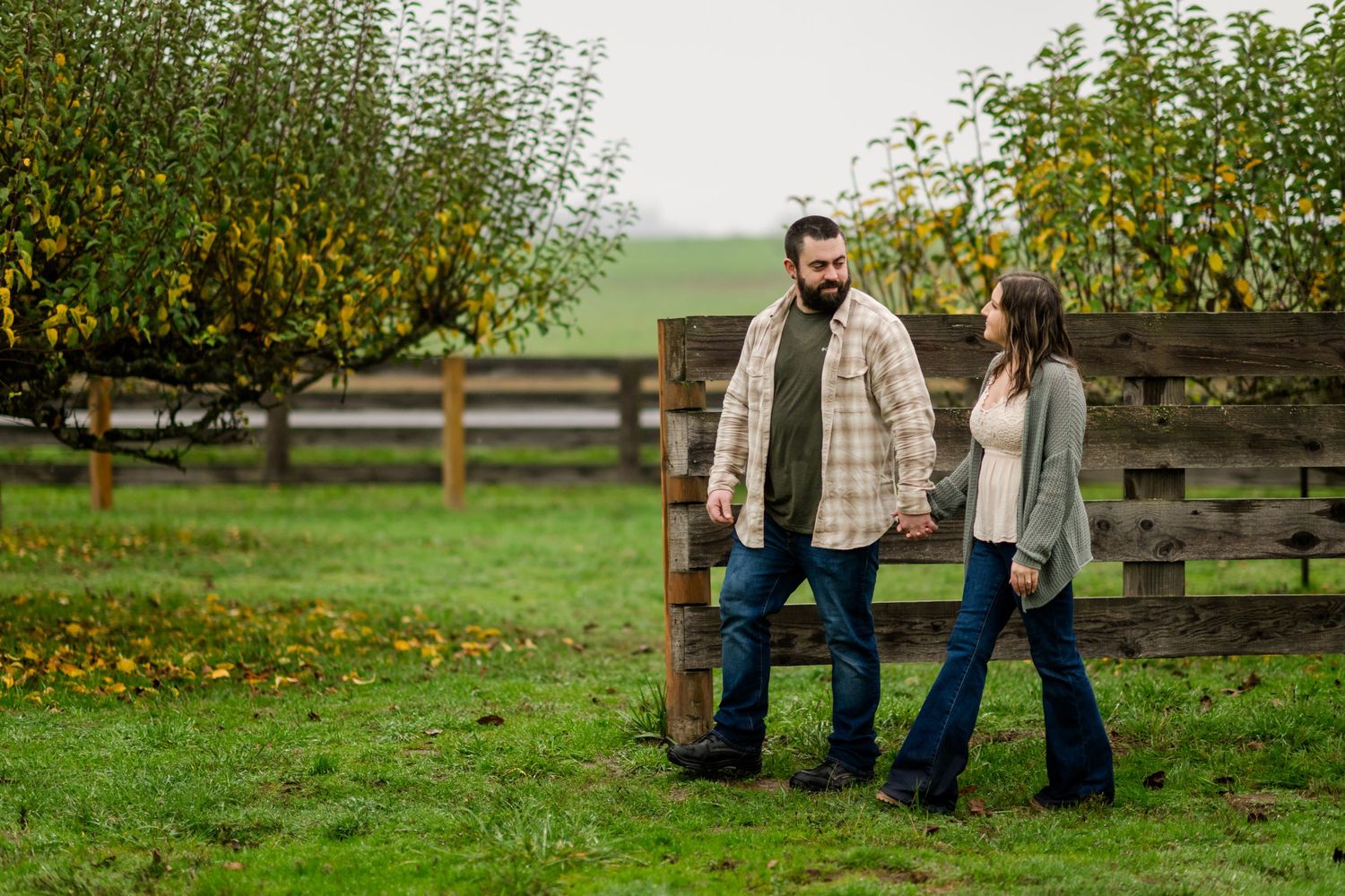 A couple holds hands while walking along a rustic wooden fence in an autumn orchard.