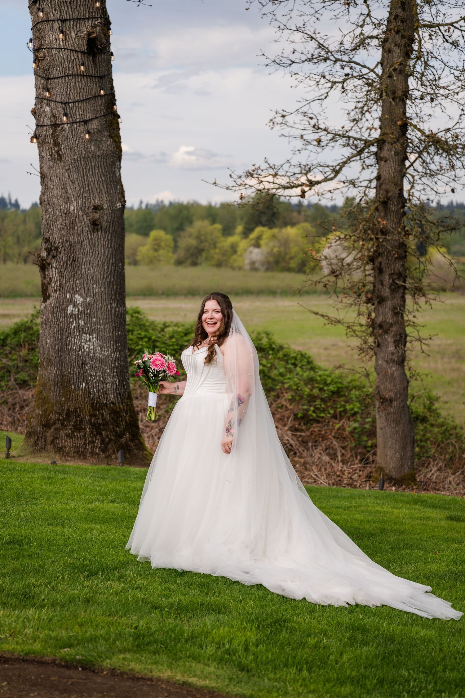 Bride in strapless ball gown with veil holds pink bouquet while standing between two trees in outdoor setting.