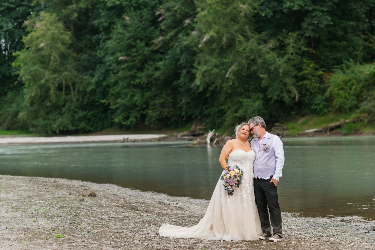Bride and groom pose by a river surrounded by lush green trees on their wedding day.
