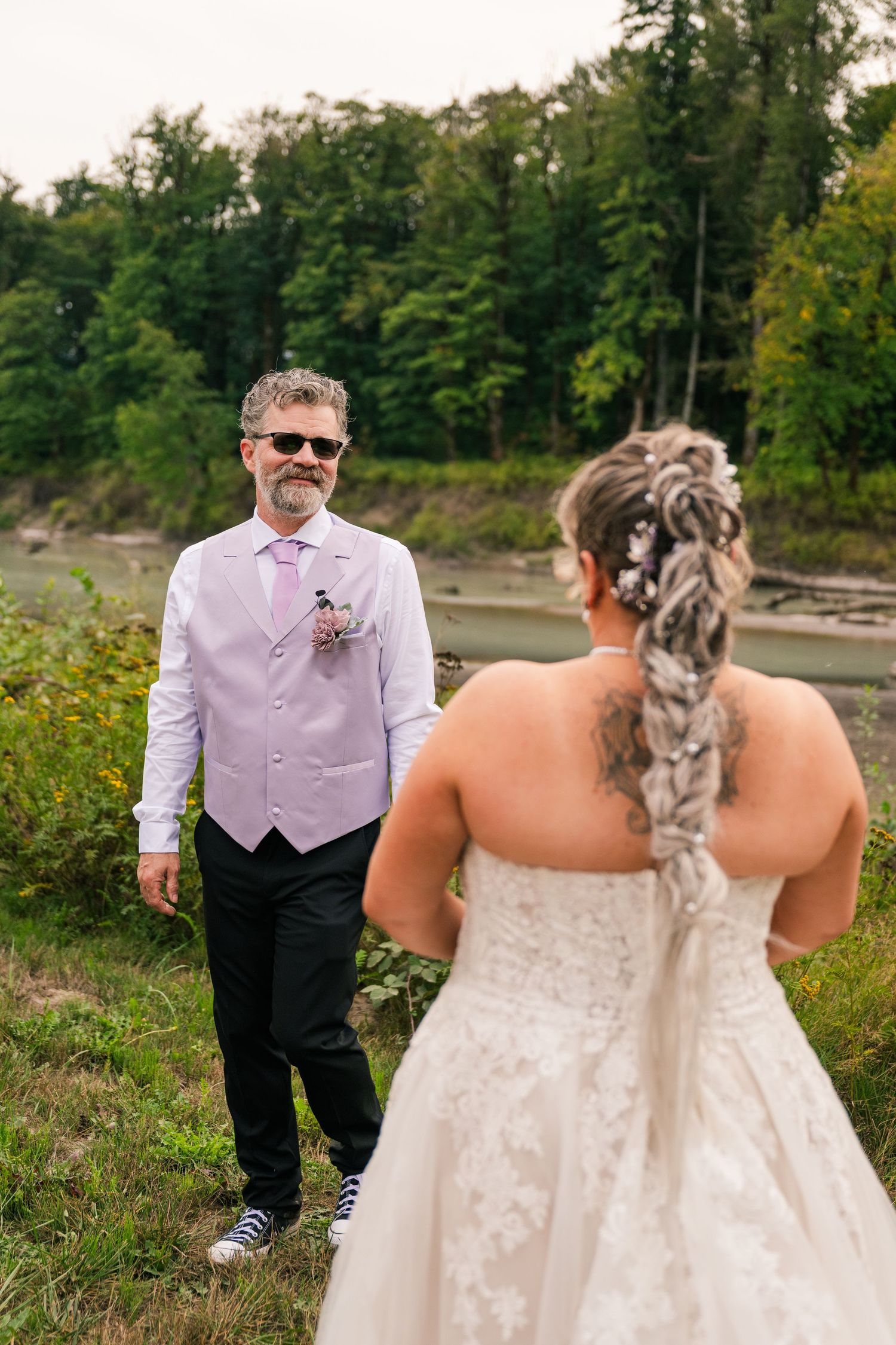 Groom in lavender vest and sunglasses watches bride in lace wedding gown during outdoor first look ceremony.